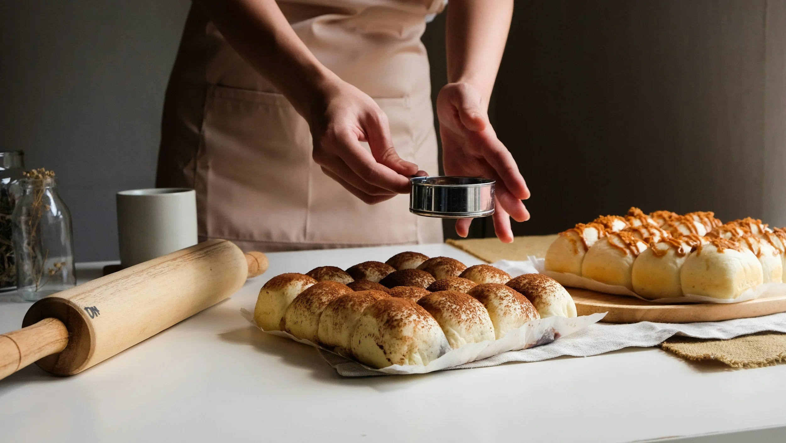 Person dusting powdered sugar on a tray of small baked goods, with another tray of cinnamon rolls topped with caramel in the background. Baking tools and ingredients are on the table.