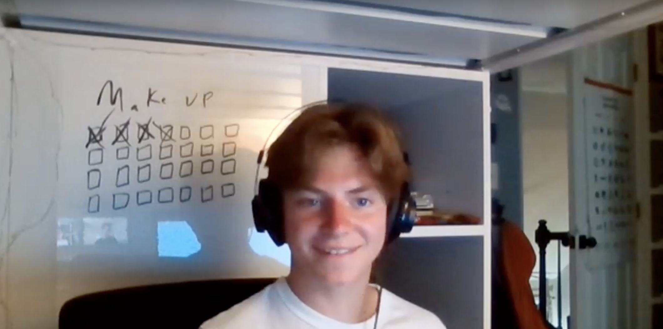 A young man with brown hair wearing headphones, smiling at the camera in a home office. Behind him, there is a whiteboard with a dotted grid with assignments written on it, and some checkmarks.