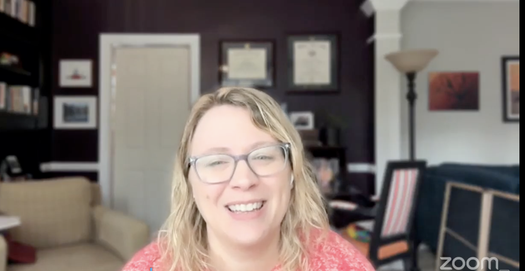 A woman with glasses and blonde hair smiling in a home office or living room with bookshelves, framed pictures, and a floor lamp in the background.