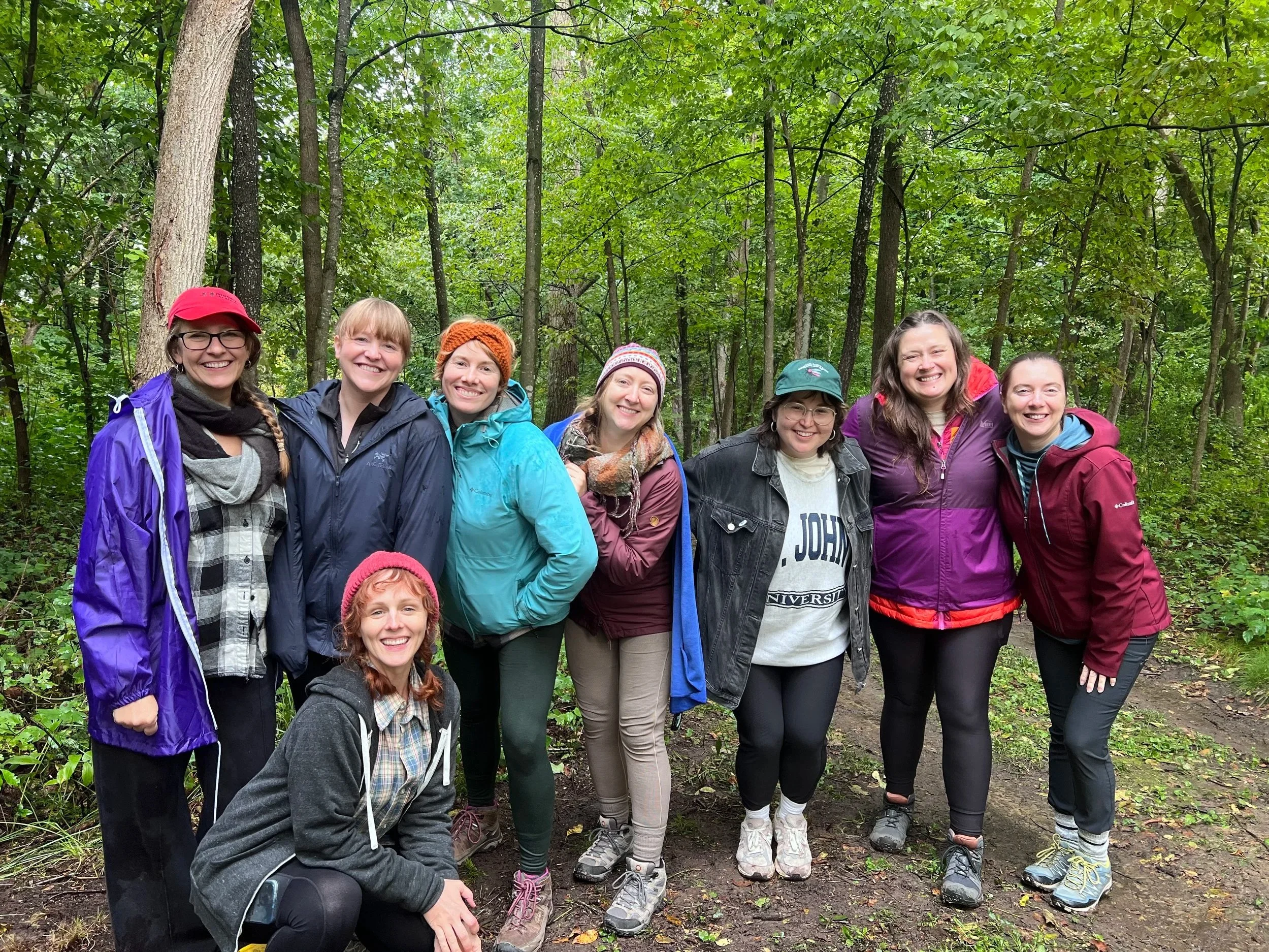 Group of nine women hiking in a forest with green trees, some wearing jackets and hats.