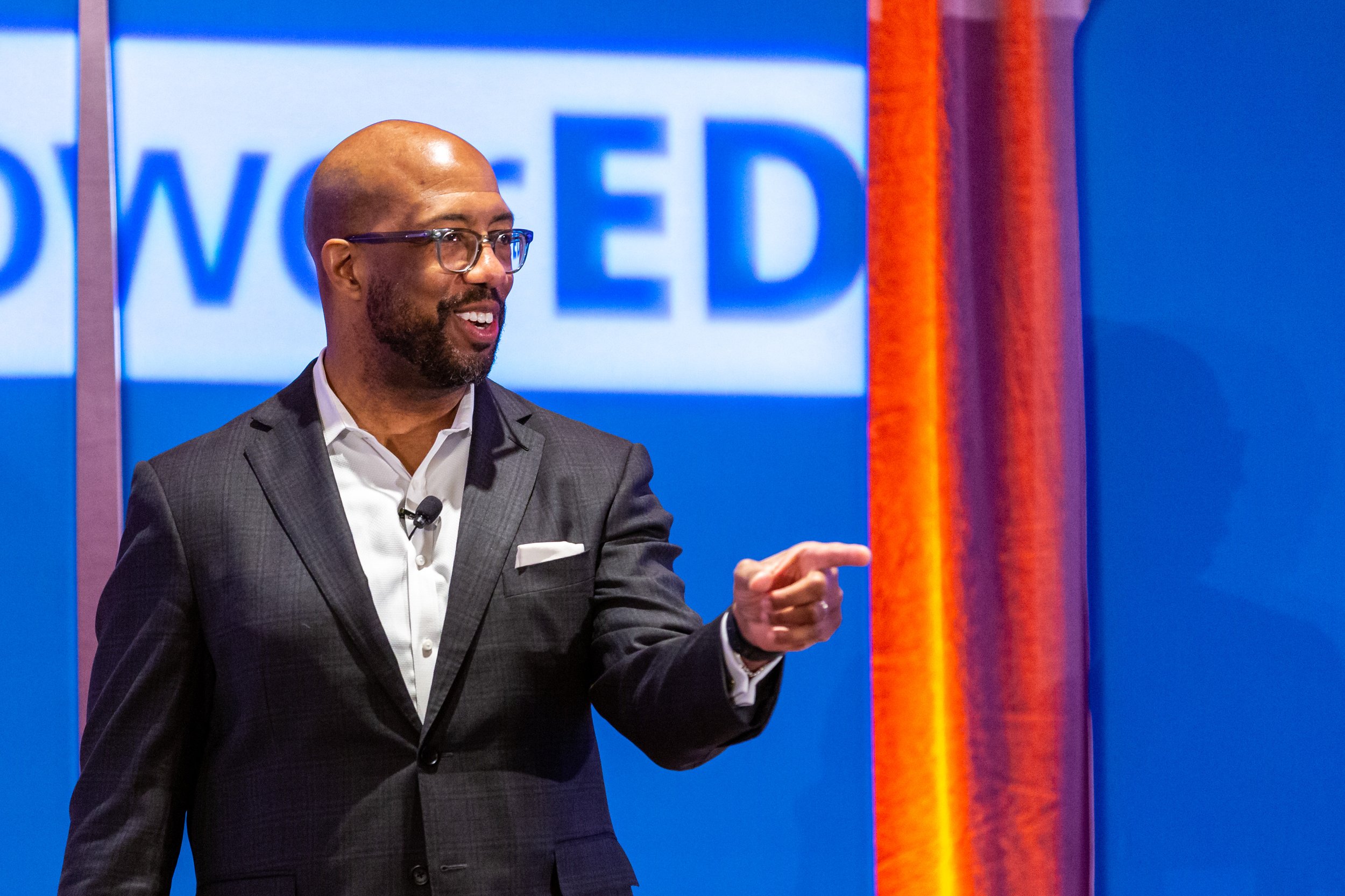 A man in a gray suit and glasses is speaking on stage with a blue background visible behind him. He is pointing forward and smiling.