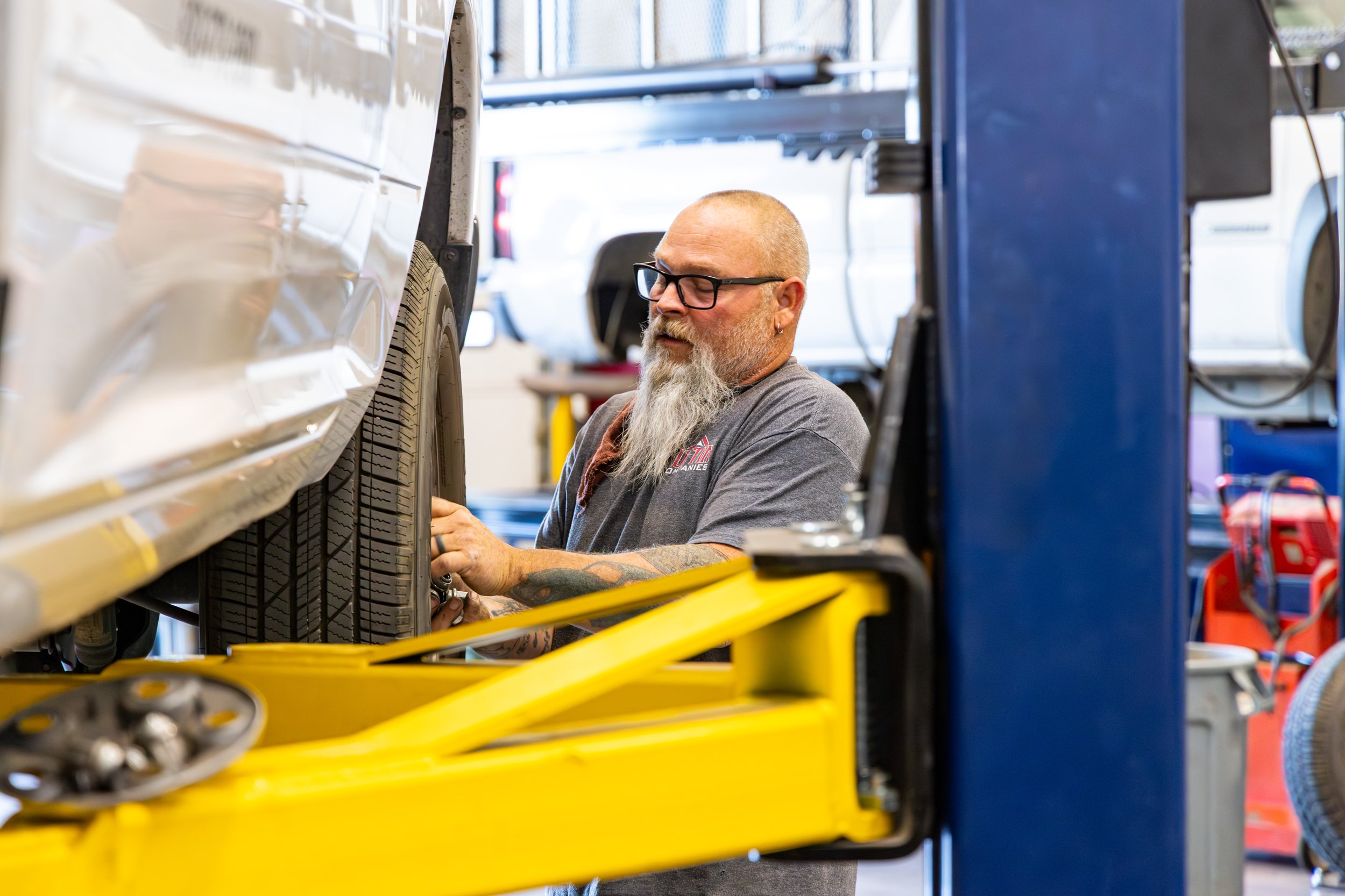 A man with glasses, a beard, and tattoos working on a vehicle's tire in an auto repair shop, standing next to a blue and yellow hydraulic lift.