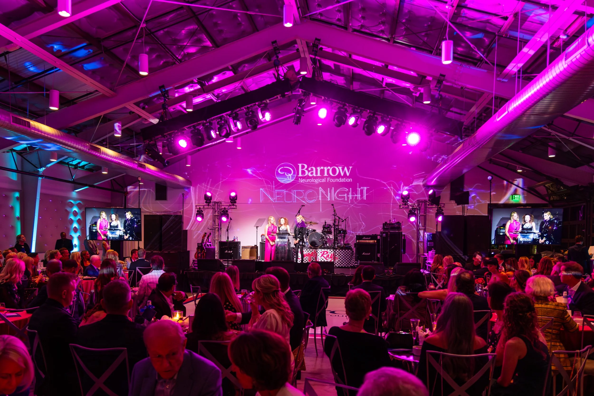 An indoor event space with a stage illuminated in pink and purple lighting where a presentation or performance is taking place. Audience members are seated at round tables, watching the stage. Large screens on either side of the stage display the event. The backdrop has the logo for Barrow Neurological Foundation and the words 'NEURO NIGHT'.