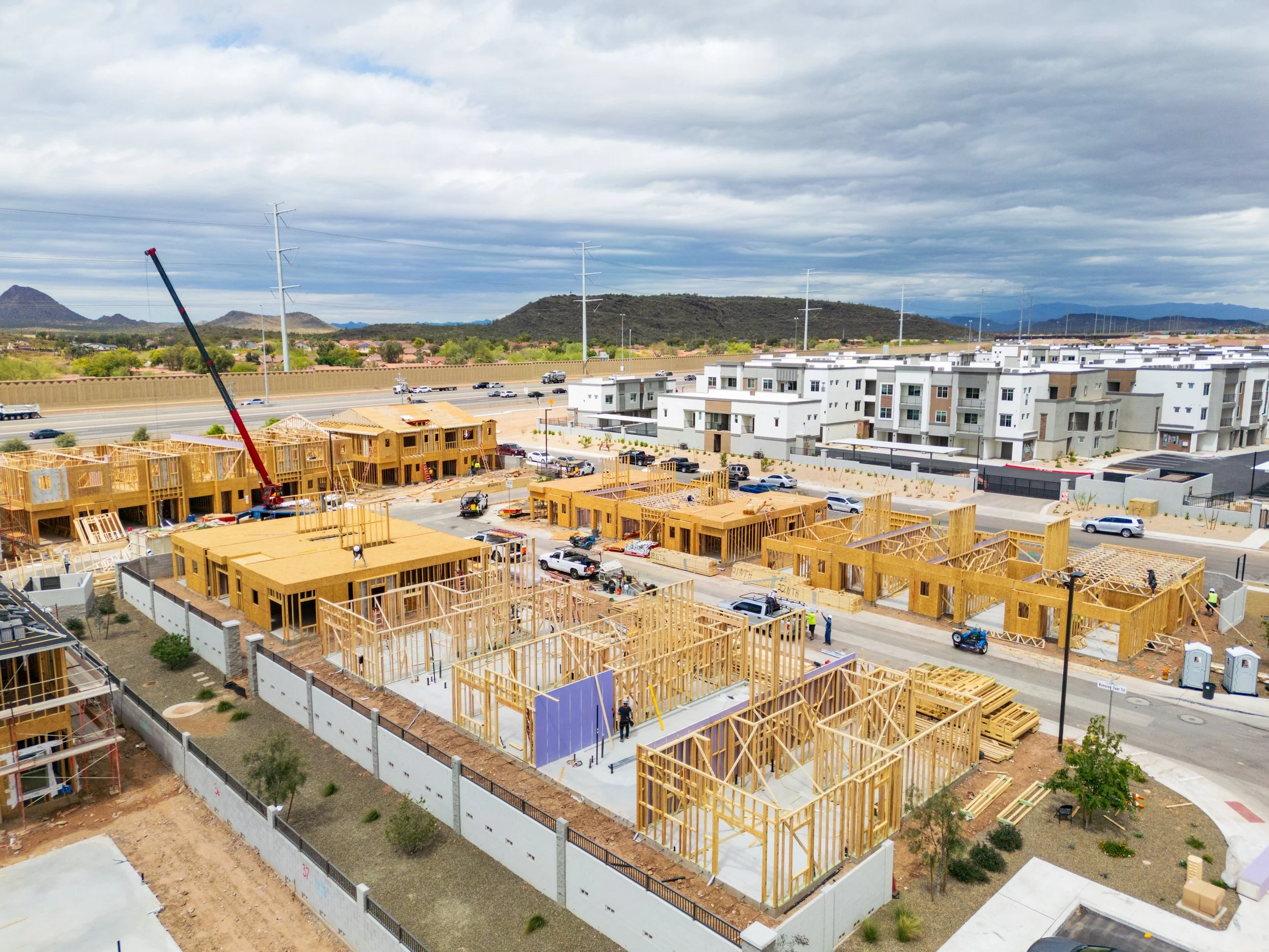 Aerial view of construction site with wooden framing for multiple upcoming houses, several workers, and construction equipment in a residential area.