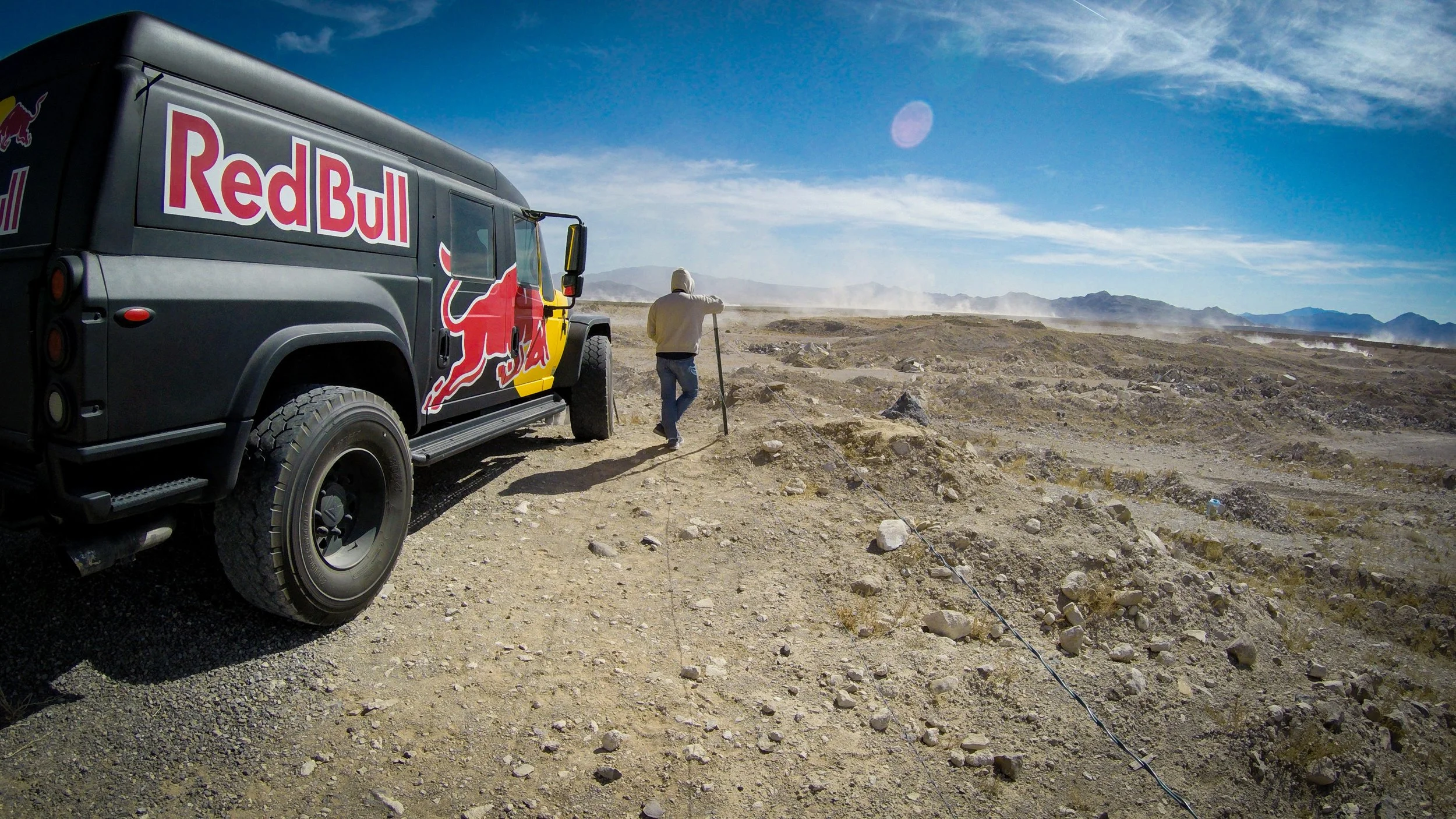 A person in a hoodie stands on rocky desert terrain near a large black vehicle with Red Bull branding, under a blue sky with wispy clouds.