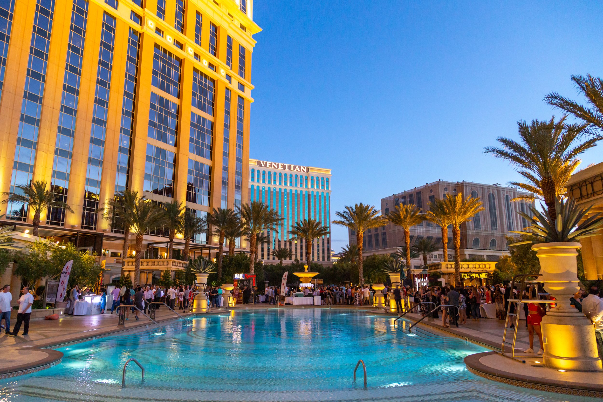Night view of a busy hotel pool area at a hotel in Las Vegas, with tall buildings, palm trees, and people mingling.