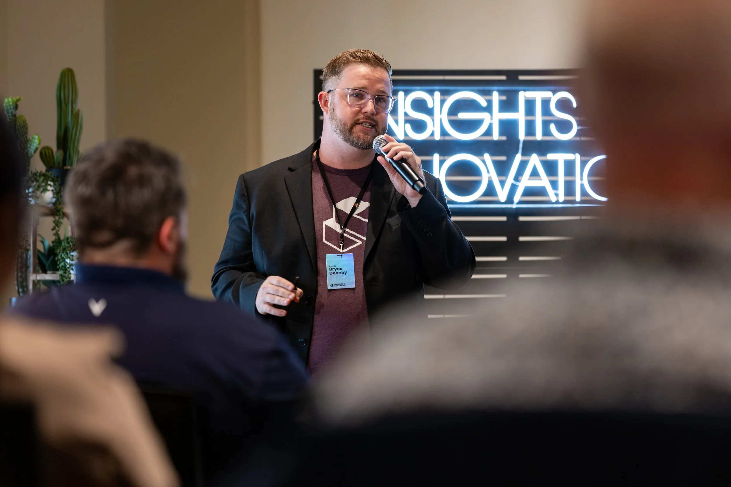 A man with glasses and a beard, wearing a black blazer and a maroon t-shirt, is giving a presentation at a conference, holding a microphone. Behind him is a neon sign that says 'INSIGHTS' and 'INNOVATION.'
