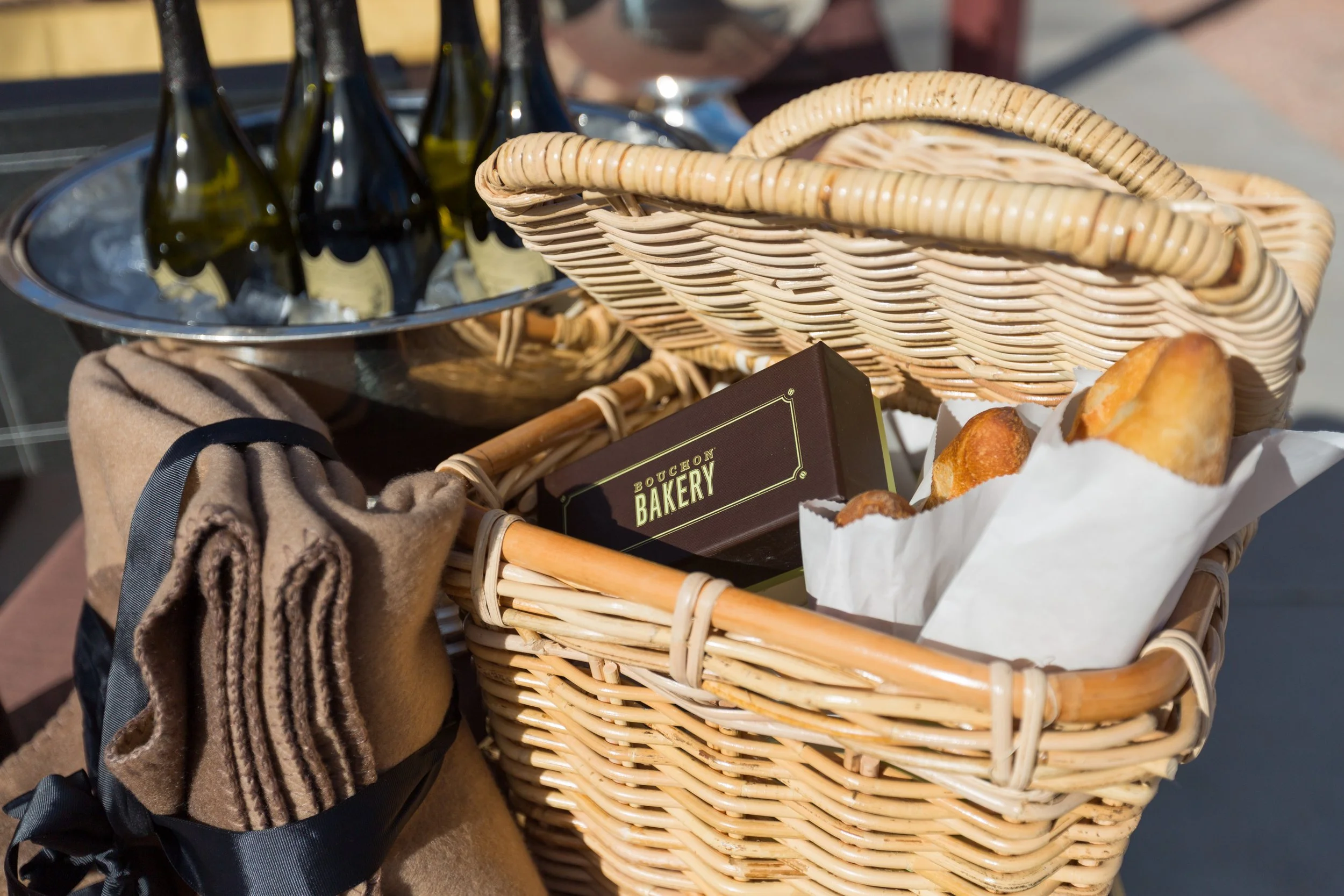 A picnic basket with bread, a box from Bouchon Bakery, a towel with a ribbon, a metal bucket with Dom Perignon champagne bottles, and a wicker container with baked goods.