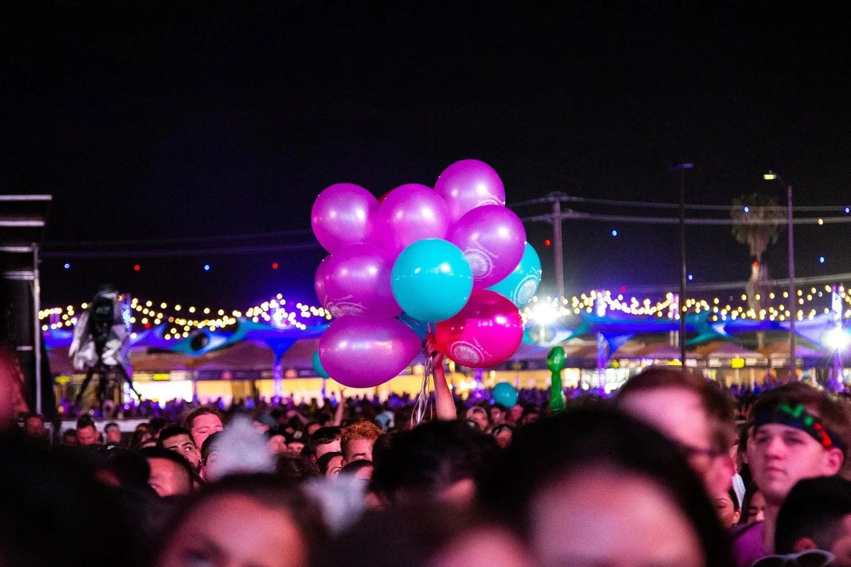 Crowd of people at night with a person holding pink, purple, and blue balloons, festival or concert setting with colorful lights and tents in the background.