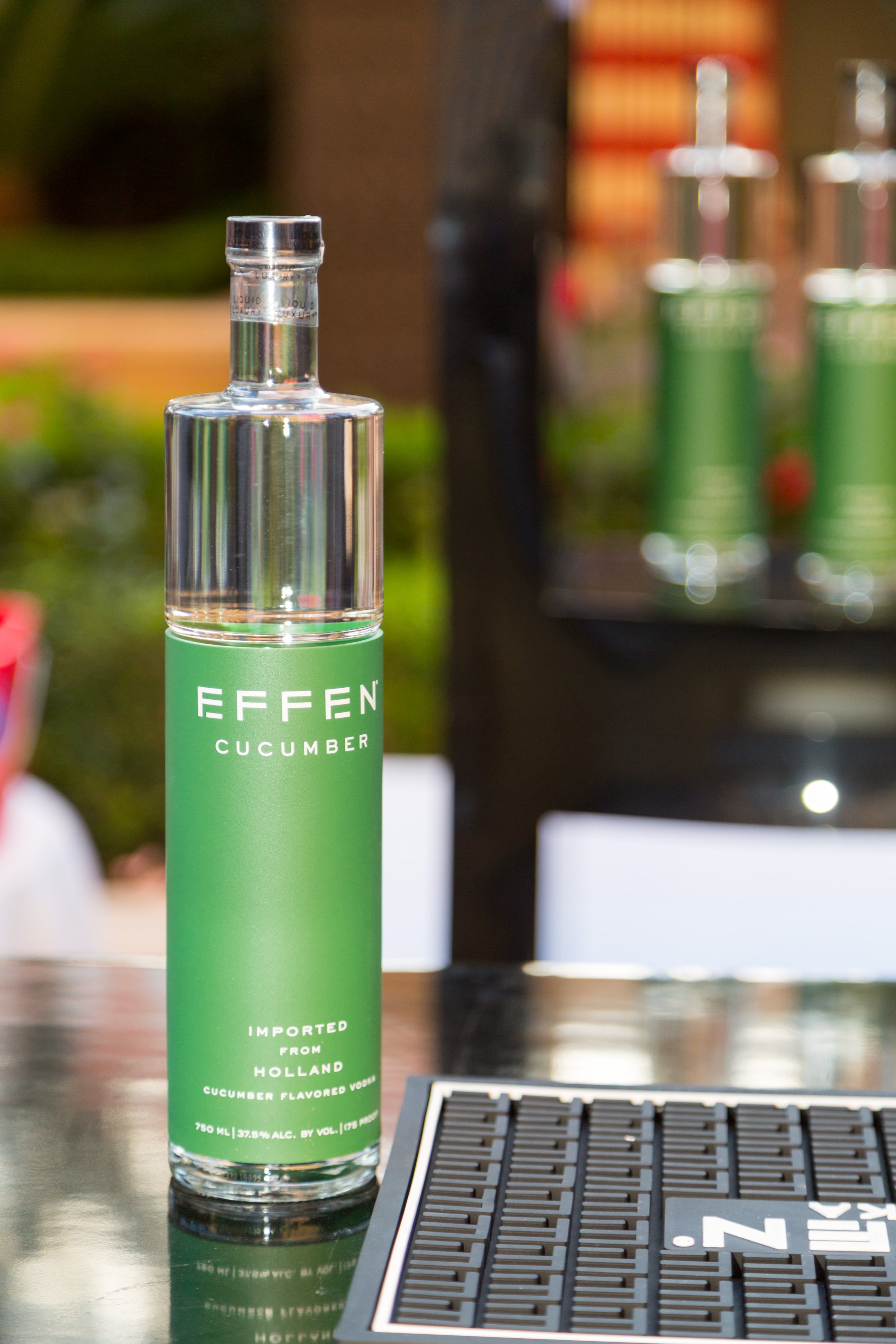 A tall glass bottle labeled 'EFFEN CUCUMBER' filled with clear liquid, placed on a reflective surface next to a black bar mat, with blurred background showing other bottles and a green outdoor setting.