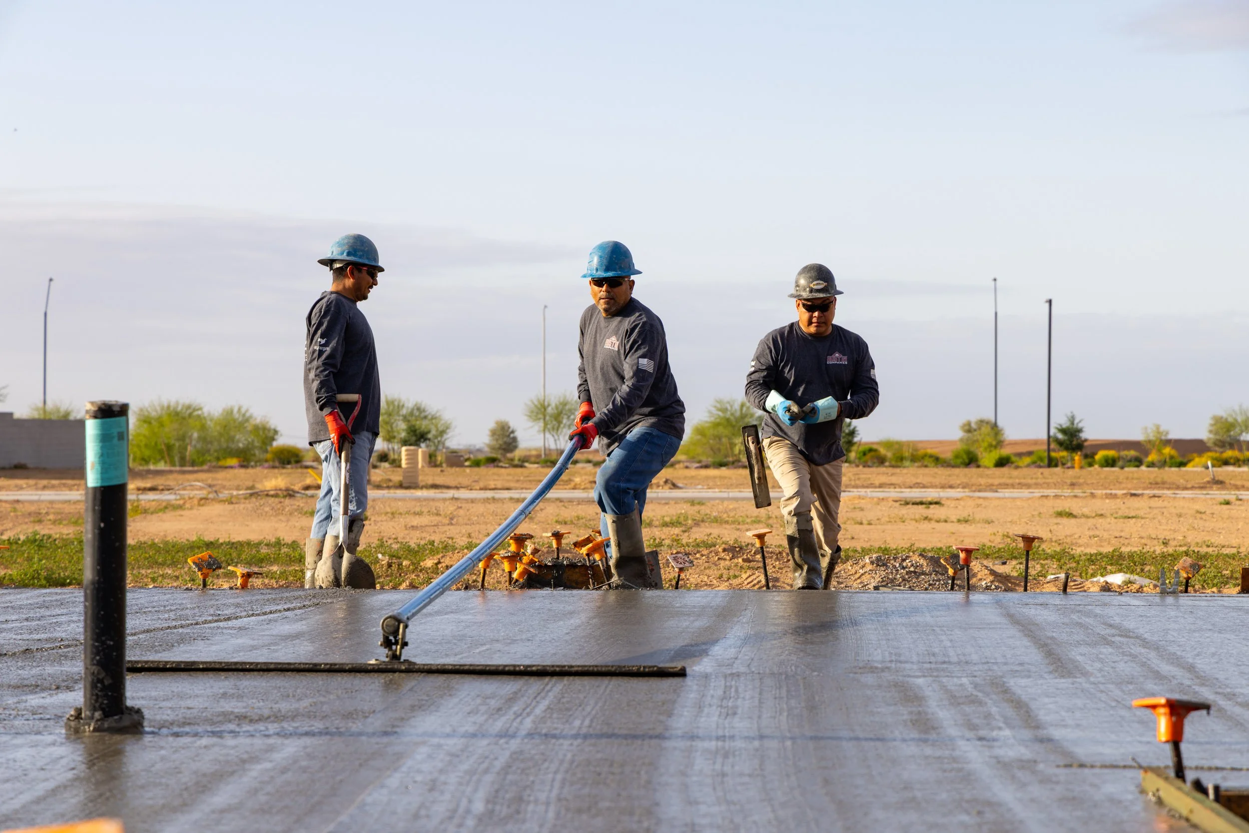 Three construction workers in safety gear pouring and spreading concrete on a flat surface at a construction site