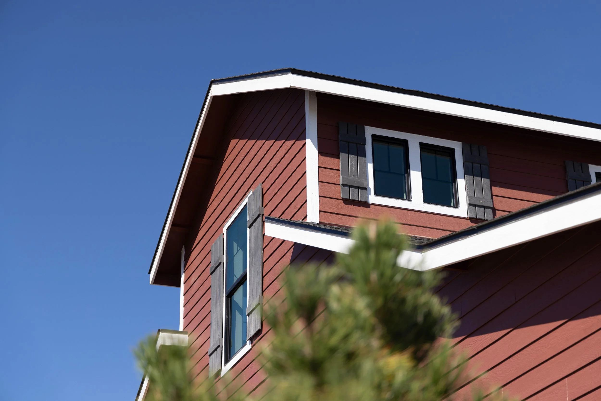 Close-up of a red house with white trim and shutters, under a clear blue sky, with a blurred green tree in the foreground.