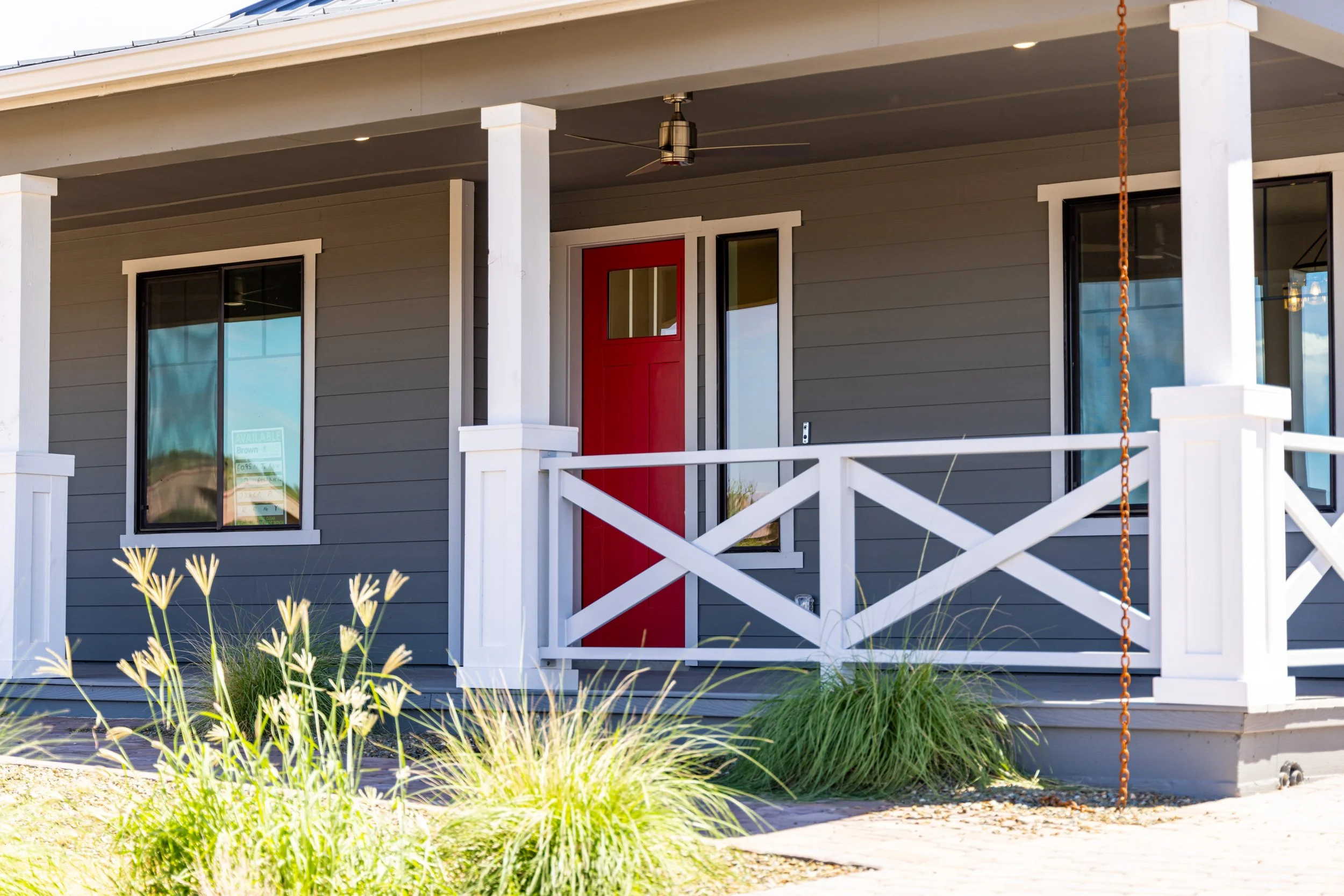 Front porch of a house with gray siding, white railing, a red front door, two windows on either side of the door, and outdoor plants in the front yard.