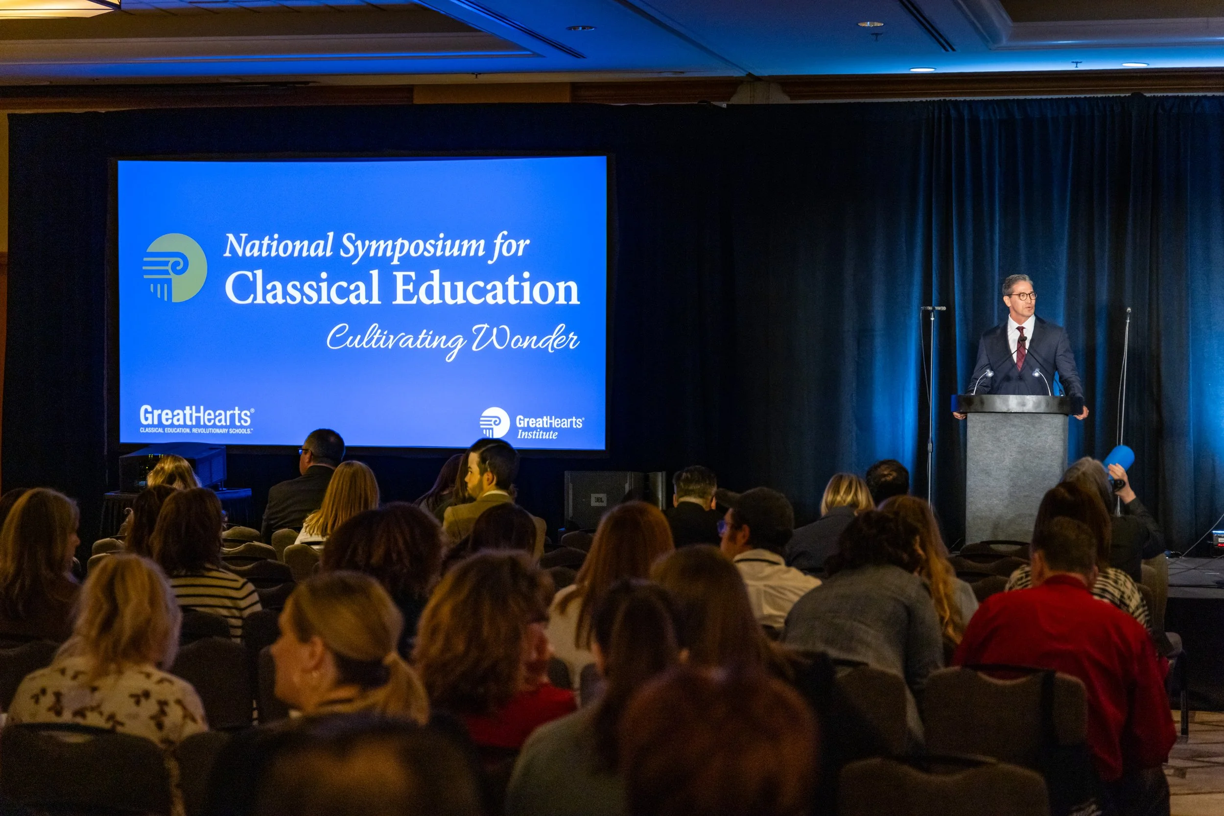 A speaker at a podium presenting at the National Symposium for Classical Education, with an audience watching. A large screen displays the event's name and tagline 'Cultivating Wonder', and the logos for Great Hearts and the Great Hearts Institute.