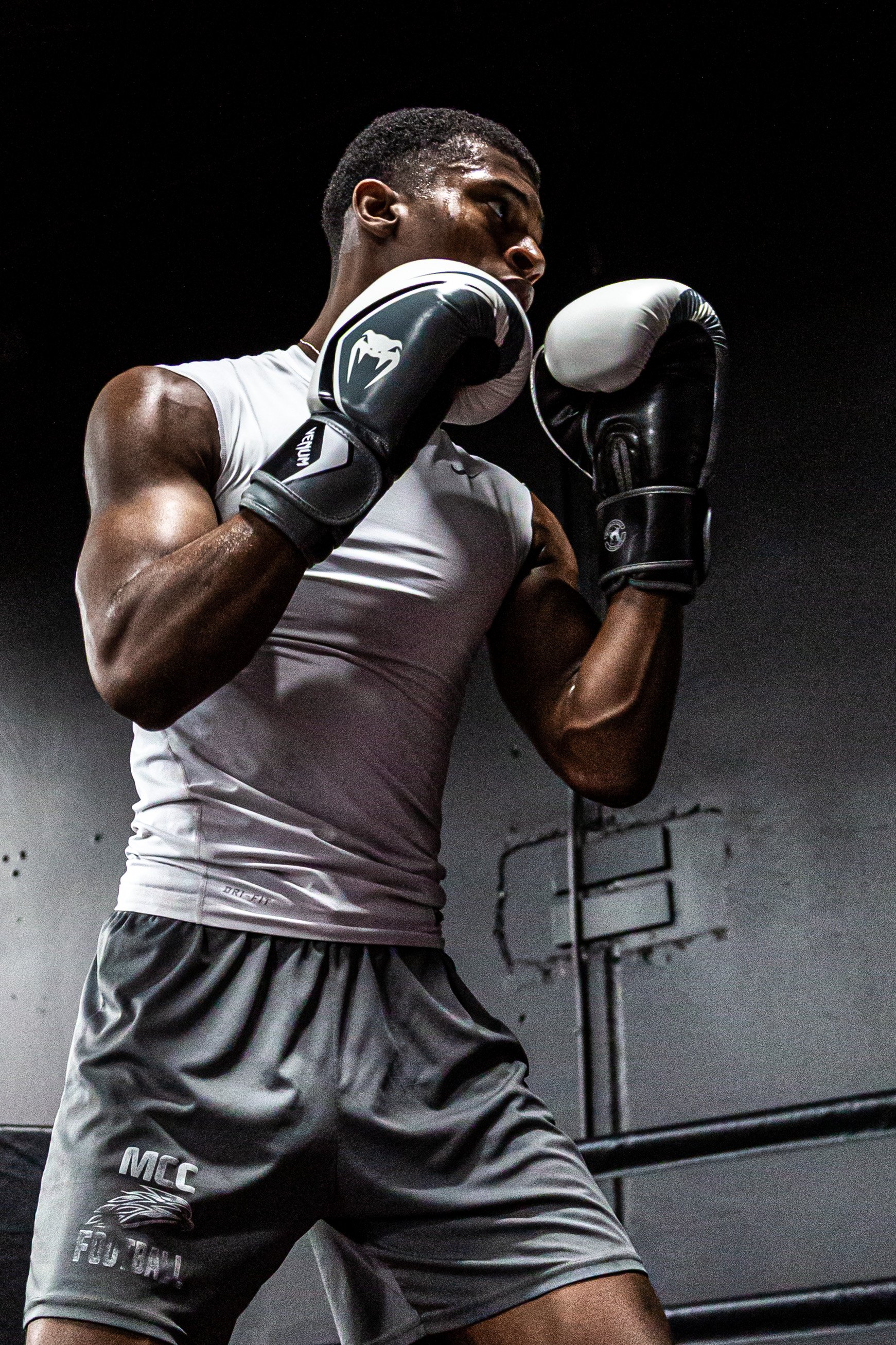A male boxer in a gym, wearing gloves and workout attire, standing in a fighting stance with fists raised near his face.