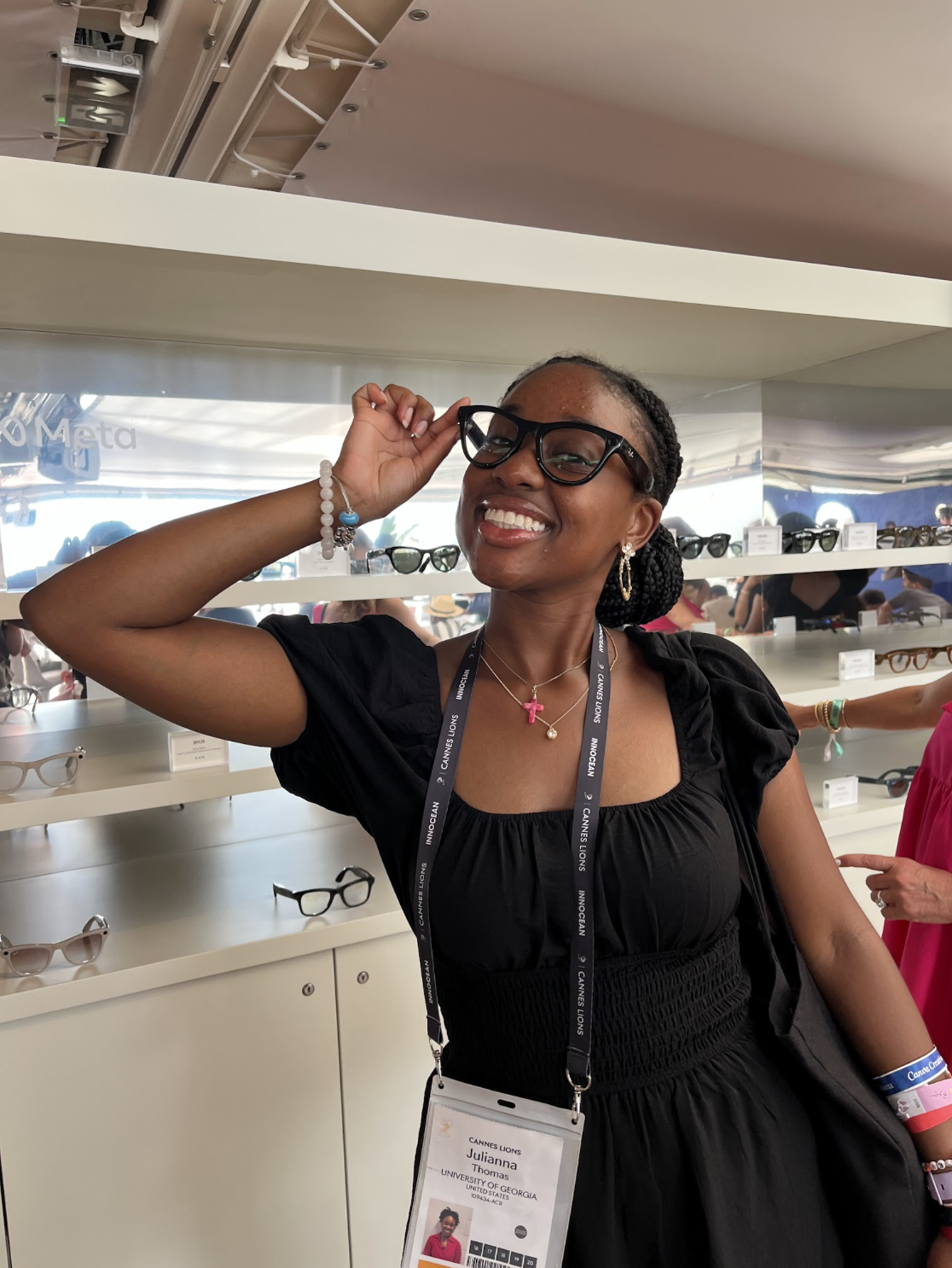 Smiling woman wearing black dress and glasses at sunglasses display, holding her glasses by the frame, with a name badge around her neck.