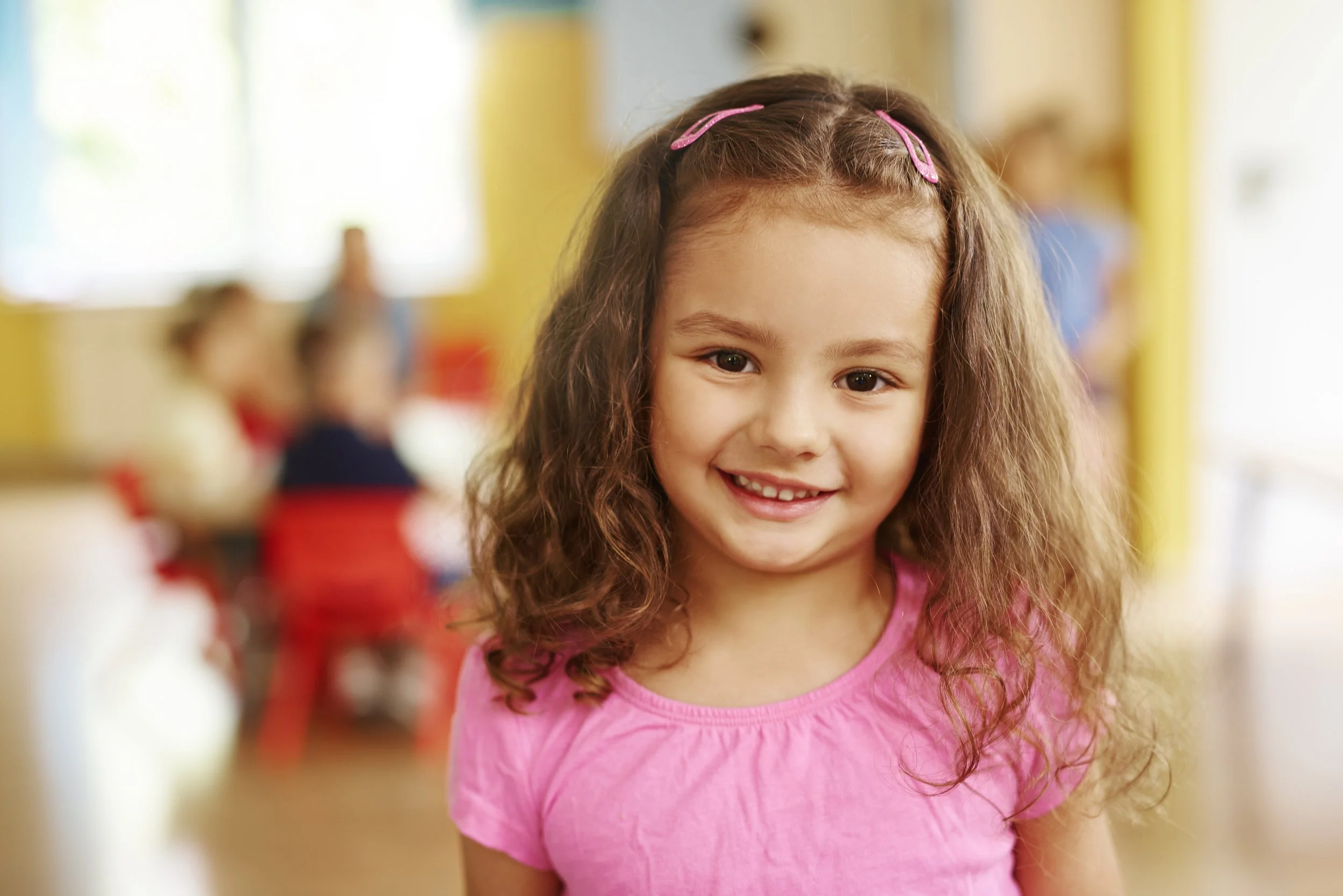 A young girl with long, curly brown hair and pink hair clips, wearing a pink shirt, smiling at the camera in a colorful indoor setting, with blurred children in the background.