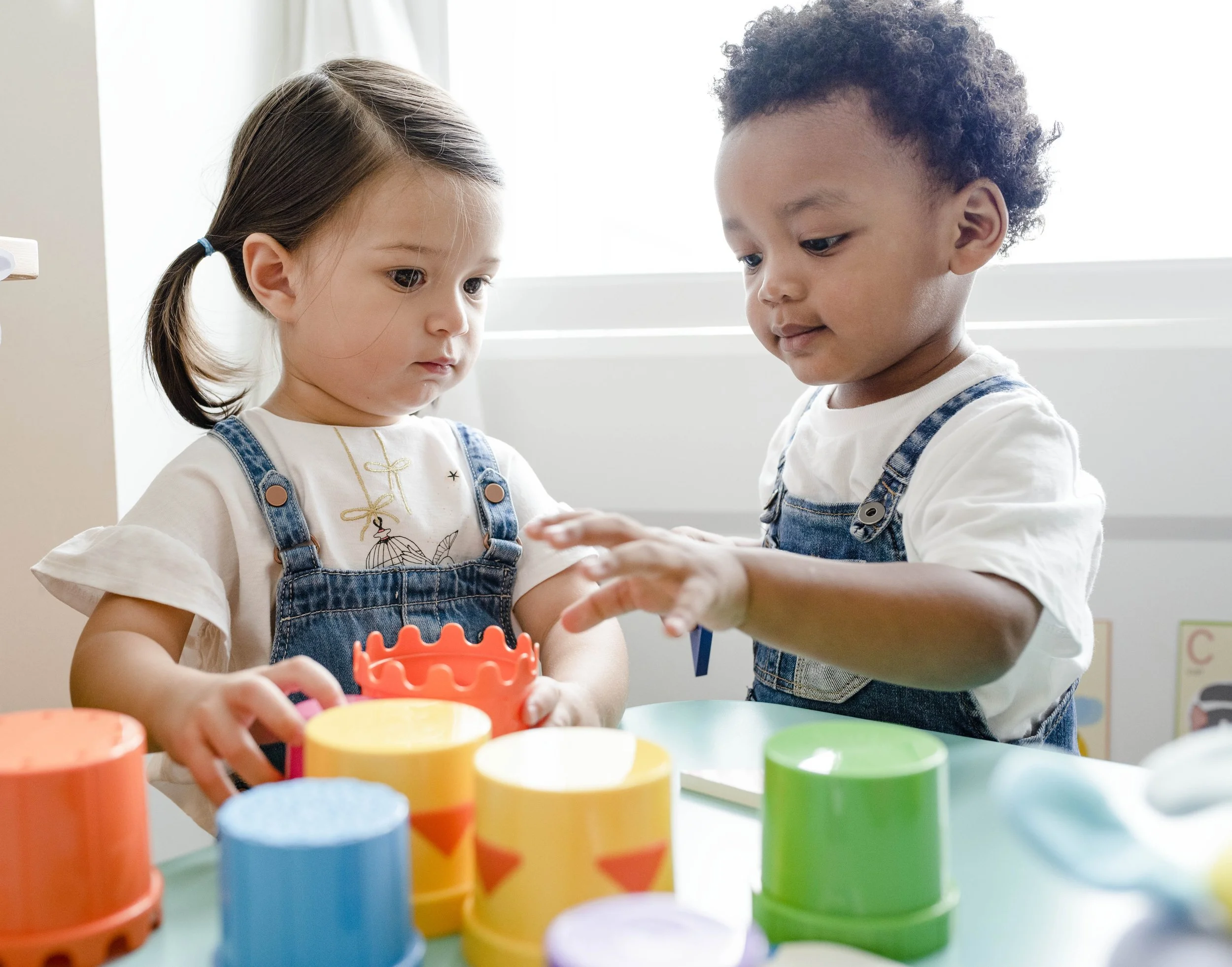 Two young children, a girl and a boy, playing with colorful stacking cups at a table in a bright room.