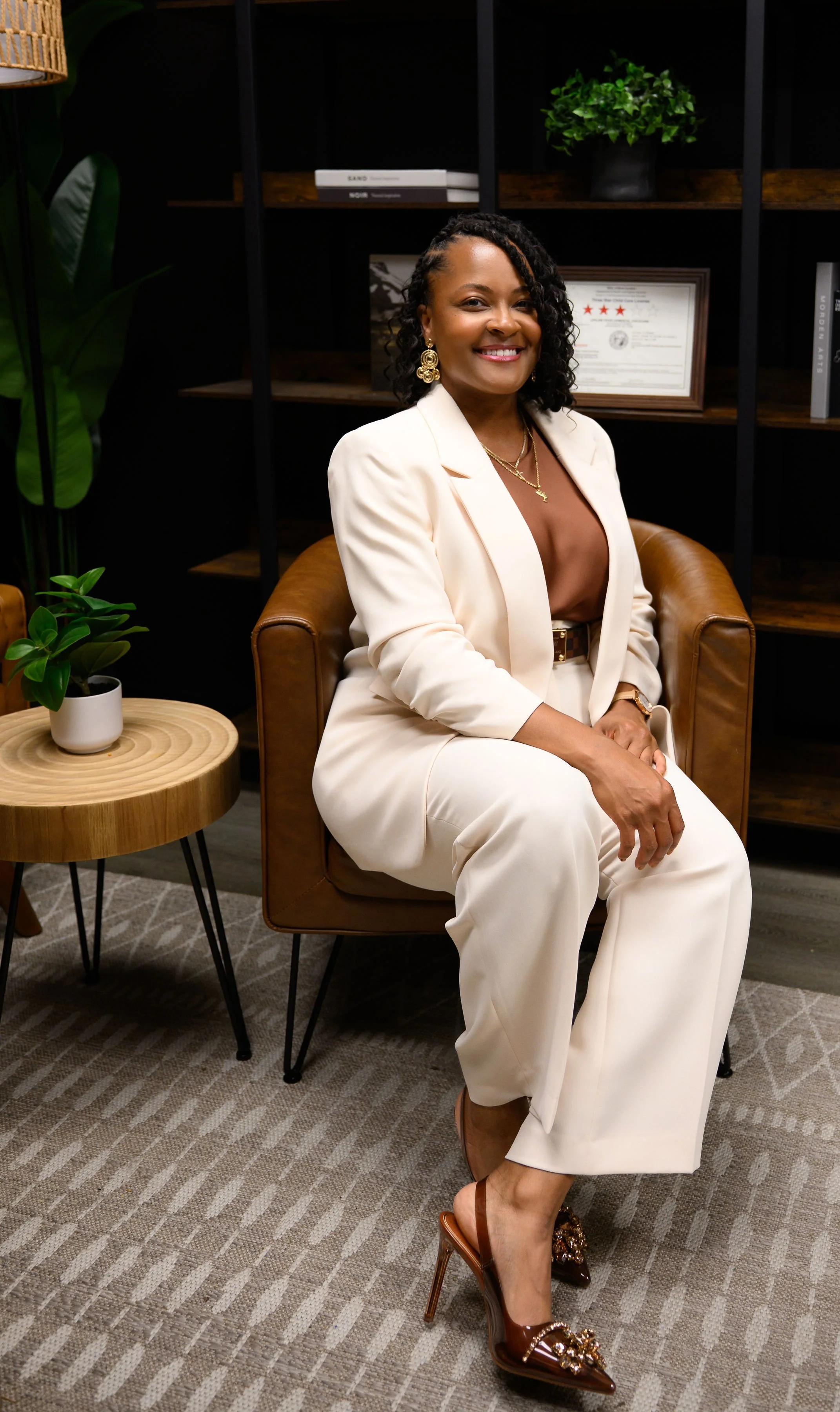 A woman in an off-white blazer and matching pants sitting in a brown leather chair in a modern office setting, smiling at the camera. She is wearing gold jewelry, including earrings and necklaces, and high-heeled shoes with embellishments.