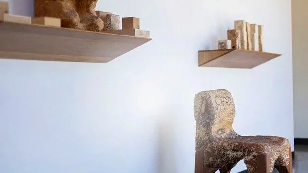 Two wooden floating shelves holding bricks of various types and sizes on a white wall, and an old, worn stone chair in the foreground.