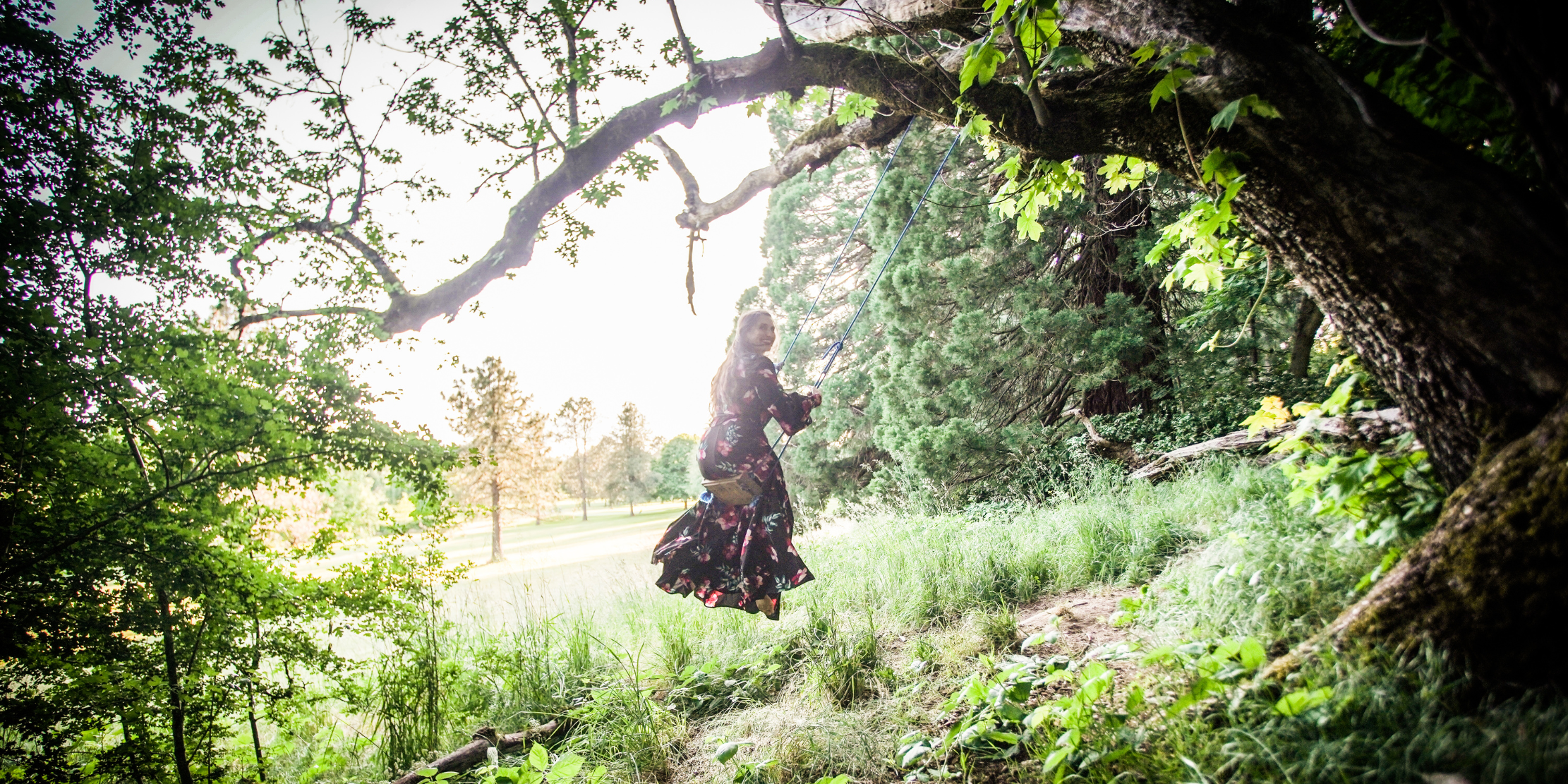 A woman swinging on a tree swing in a lush green park or forest, with sunlight filtering through the trees.