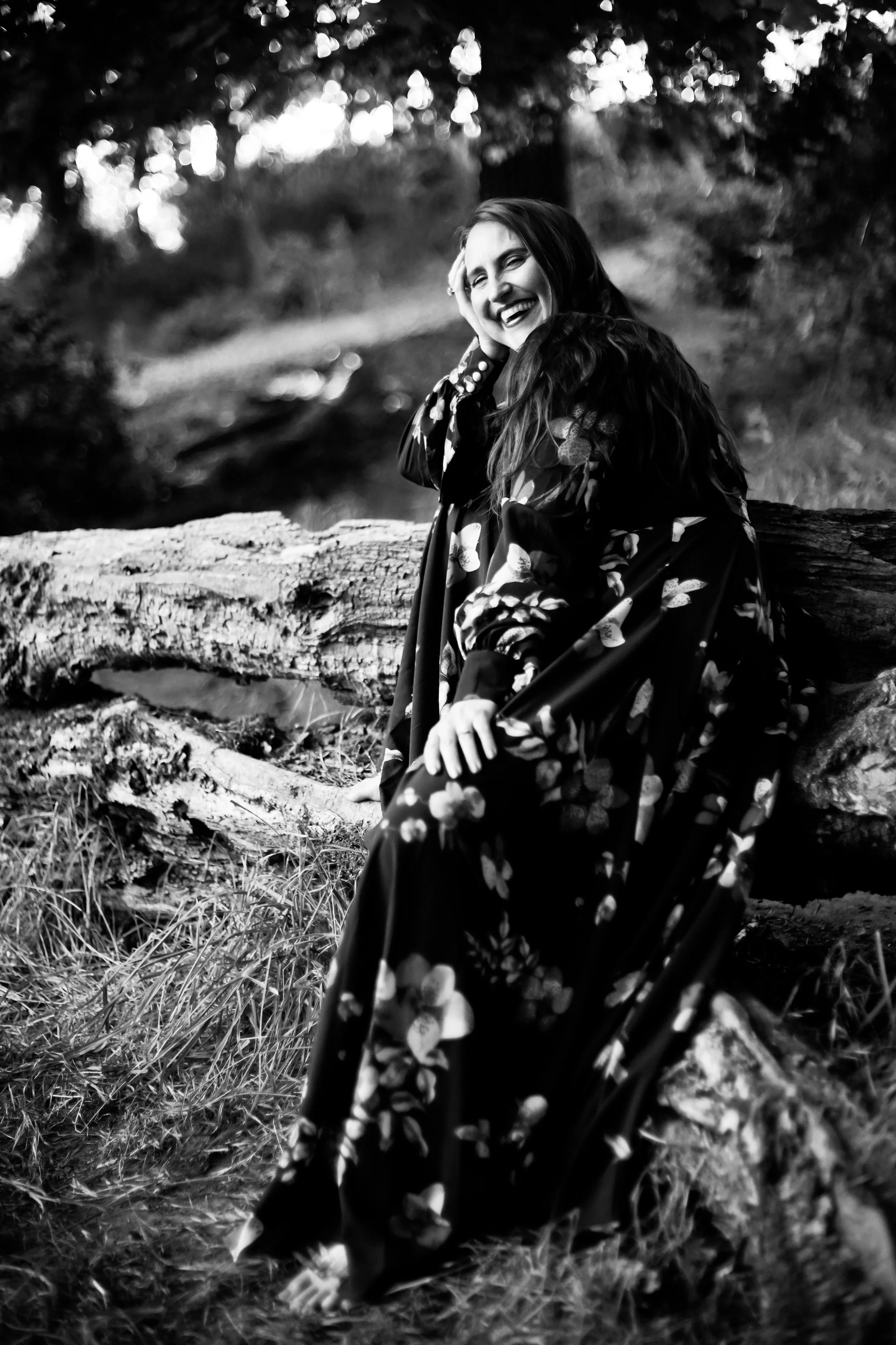 A woman in a floral dress sitting on a fallen log in a wooded outdoor setting, smiling and laughing.