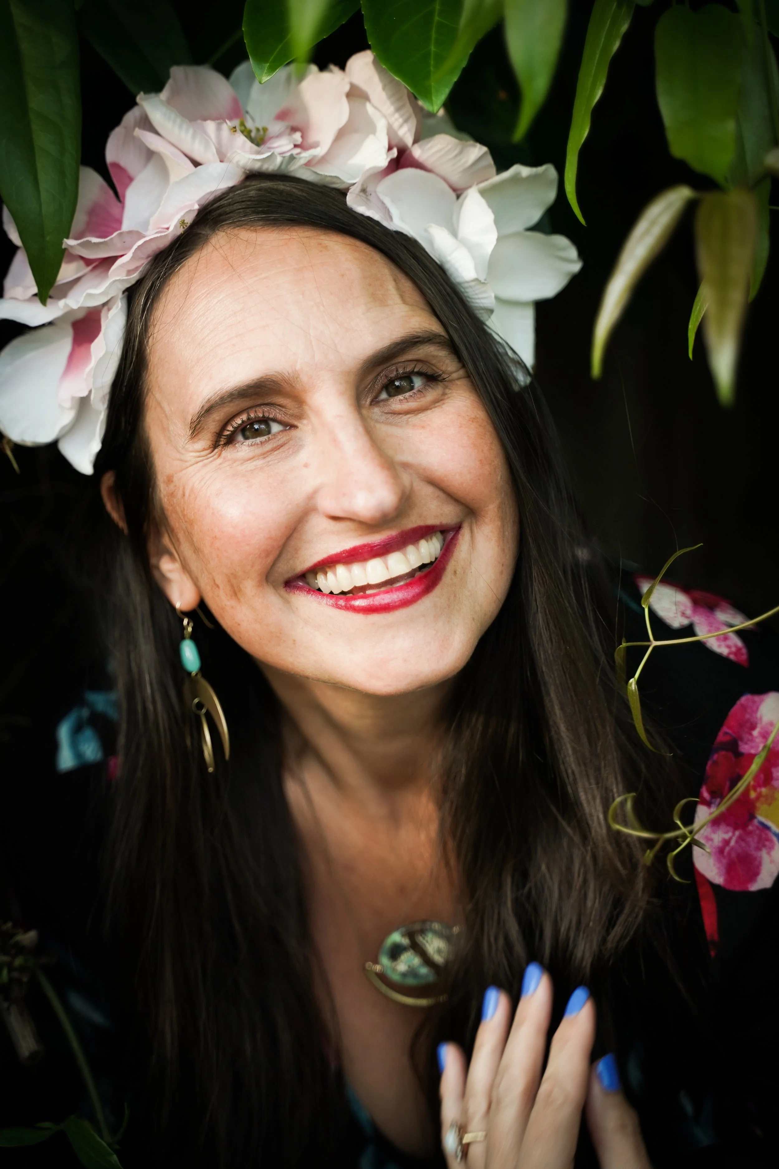 A smiling woman with long dark hair wearing a floral headpiece, earrings, and a necklace, surrounded by green leaves and flowers.