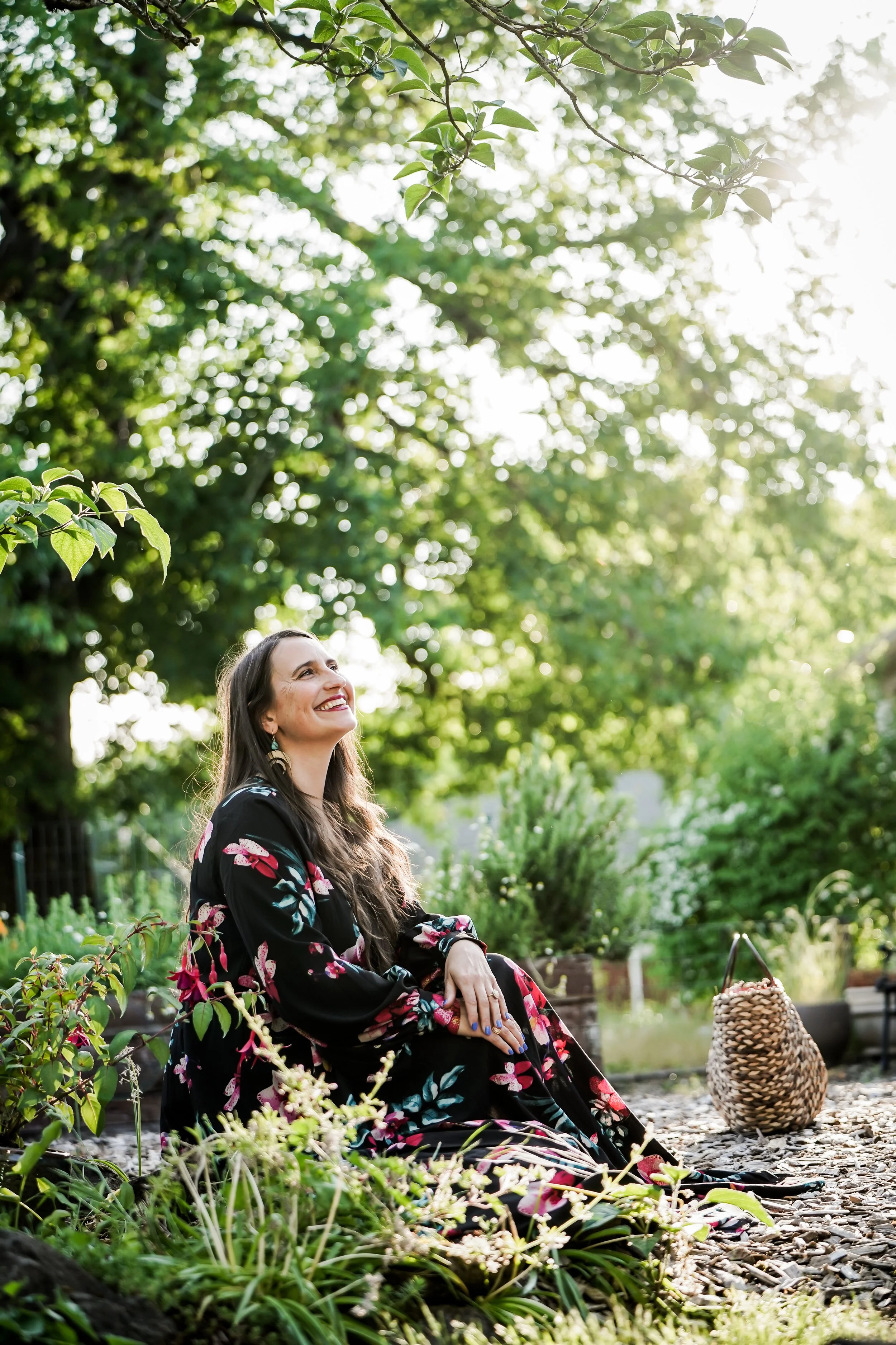 A woman in a black floral dress sitting on the ground in a garden, smiling and looking up at the sunlight filtering through trees.