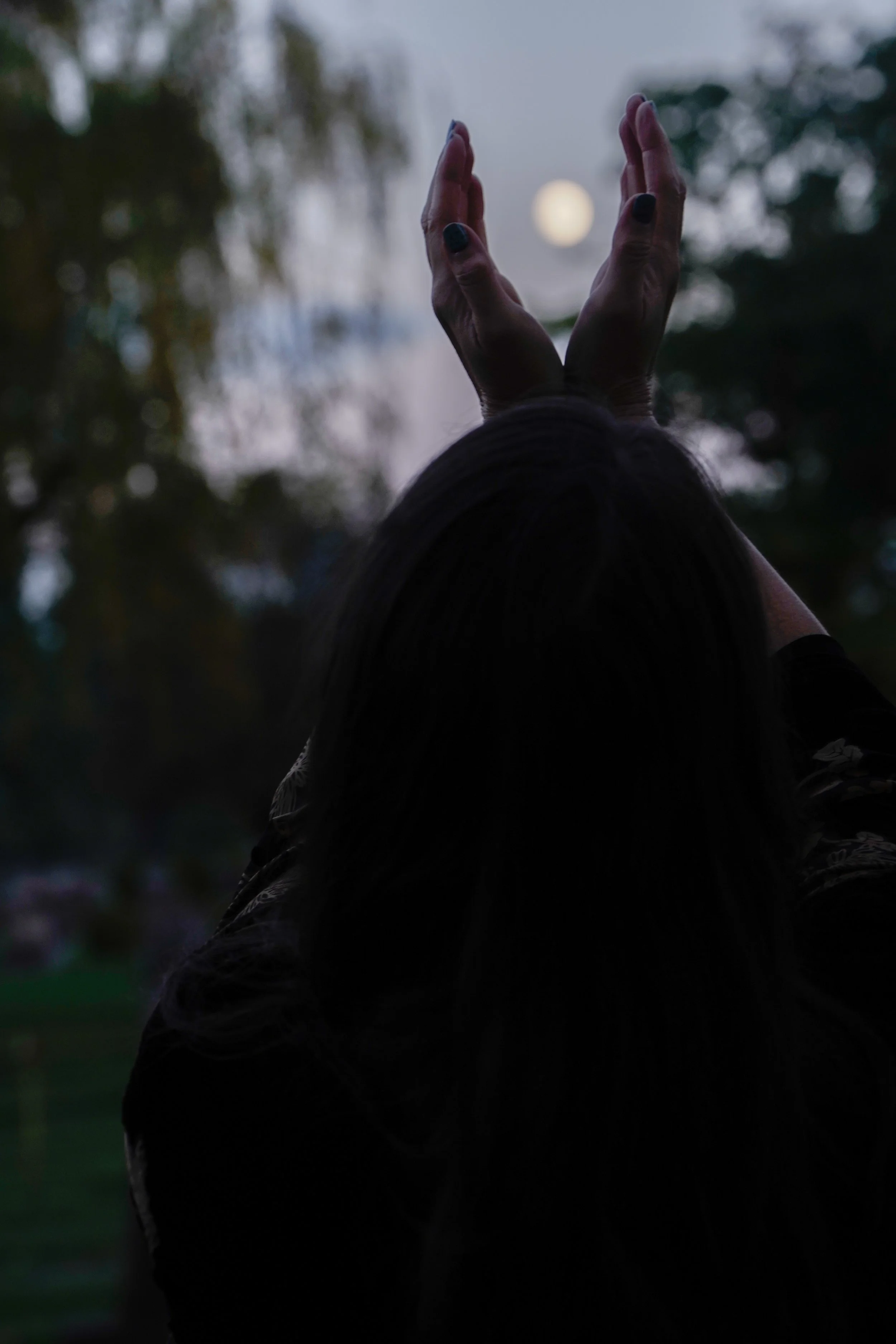 Person with dark hair holding hands up towards a full moon in a dark outdoor setting with trees in the background.