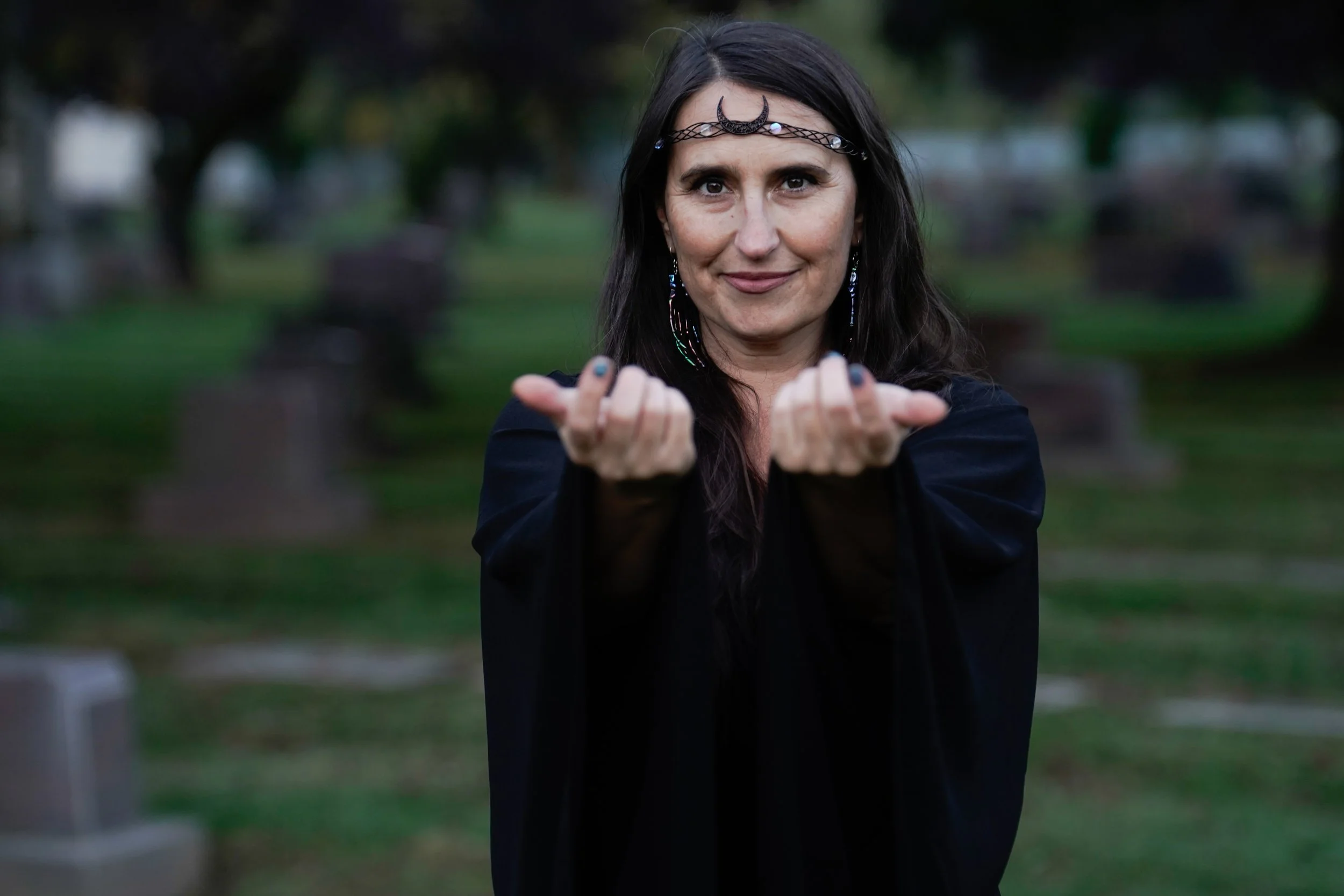 A woman with long dark hair, wearing a headband and black clothing, stands outdoors in a park with green grass and trees, making a gesture with both hands pointing towards the camera with index fingers.