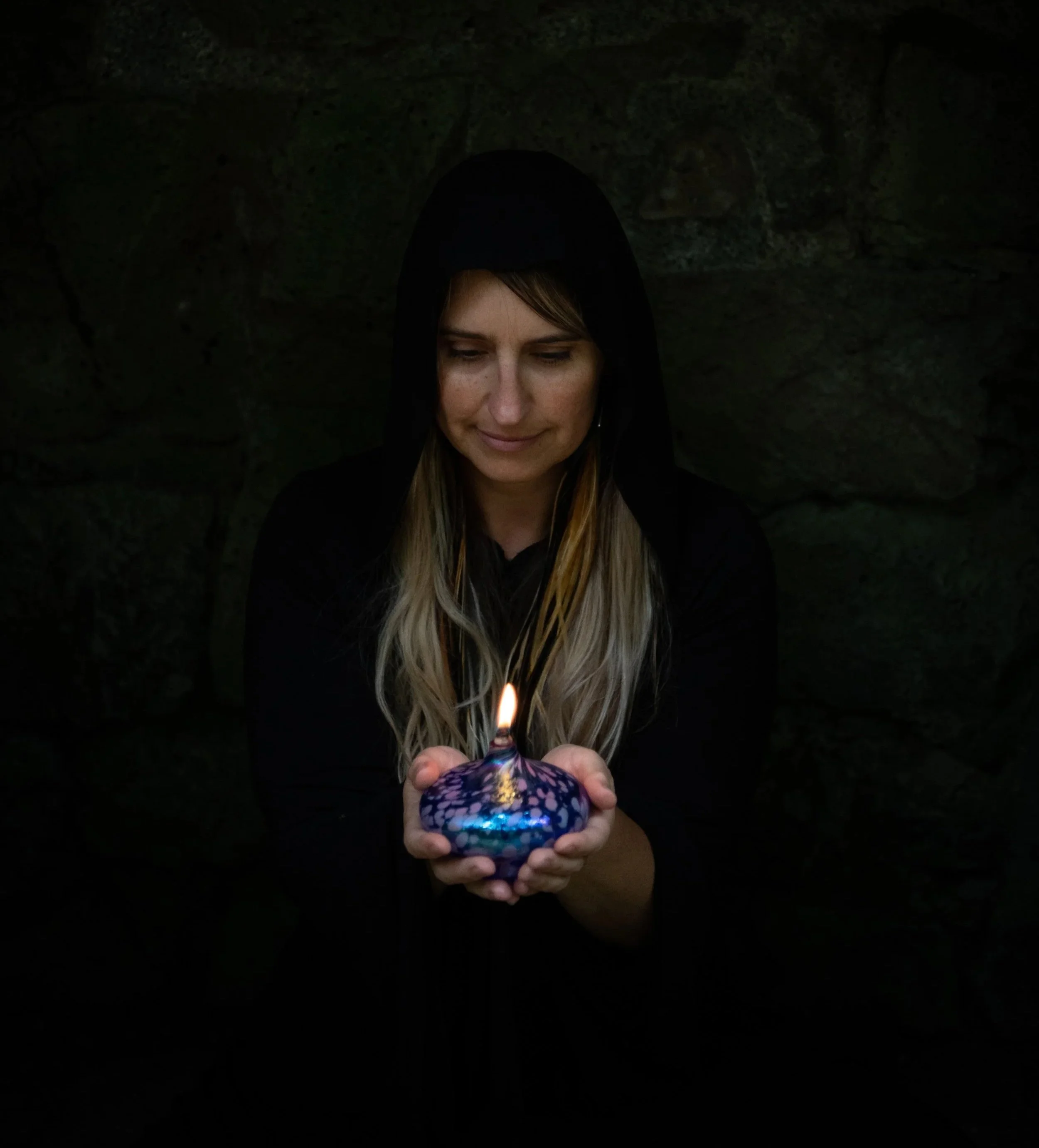 Woman holding a lit candle in a dark room or cave, wearing a black hoodie, smiling softly.