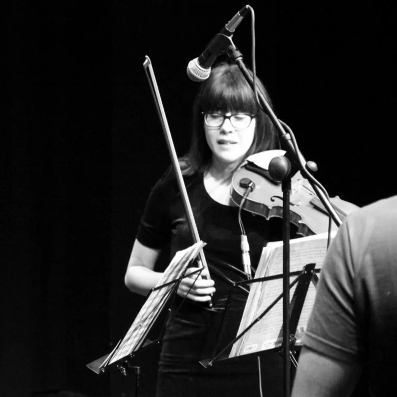 A woman with glasses and dark hair is playing a violin during a recording session, with sheet music in front of her and a microphone overhead. A sound engineer shoulder appears at the corner.