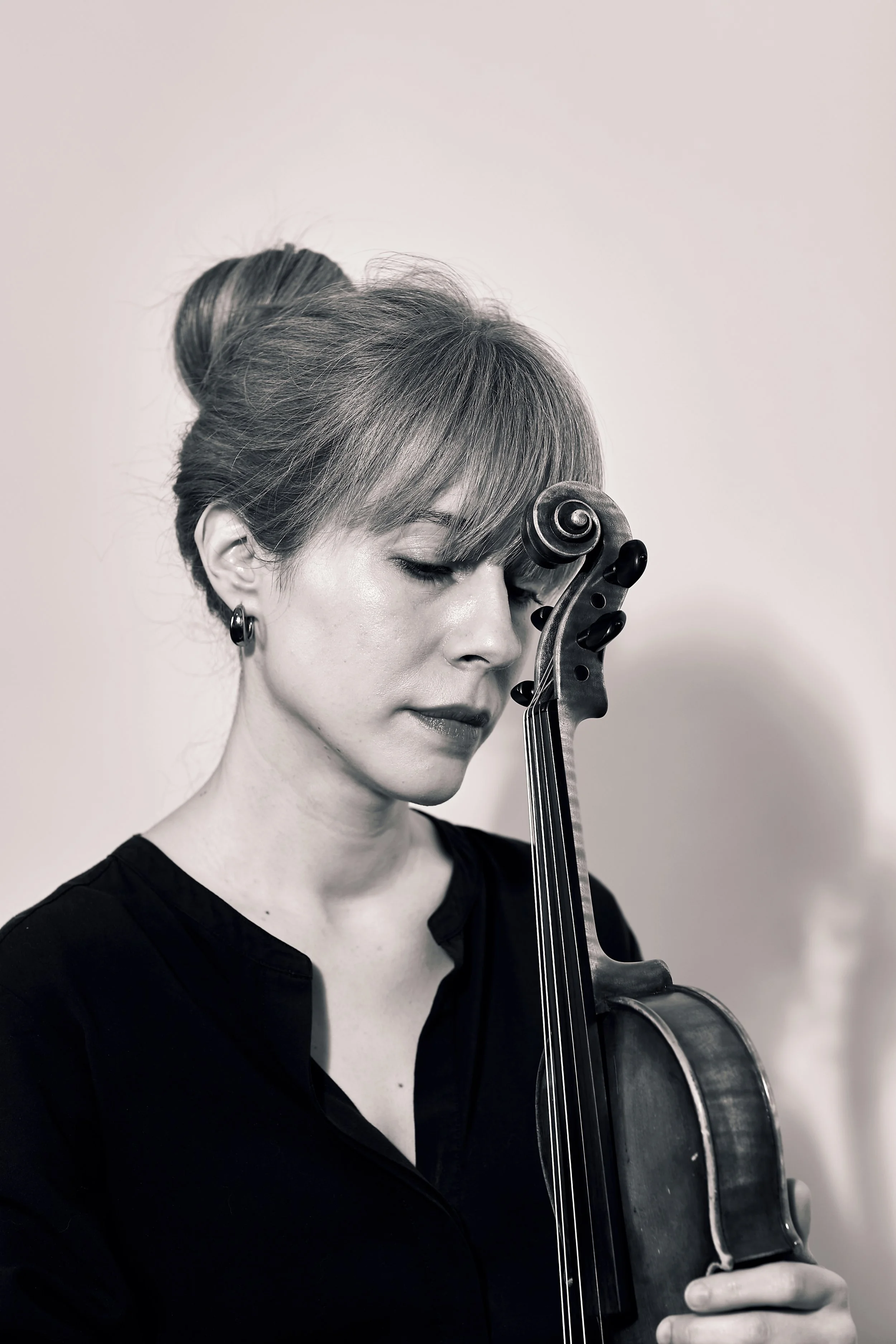Black-and-white photograph of a woman, Talia Ramos, with light-coloured hair in a bun, wearing earrings and a dark top, holding a violin close to her face, eyes closed, in a peaceful pose.