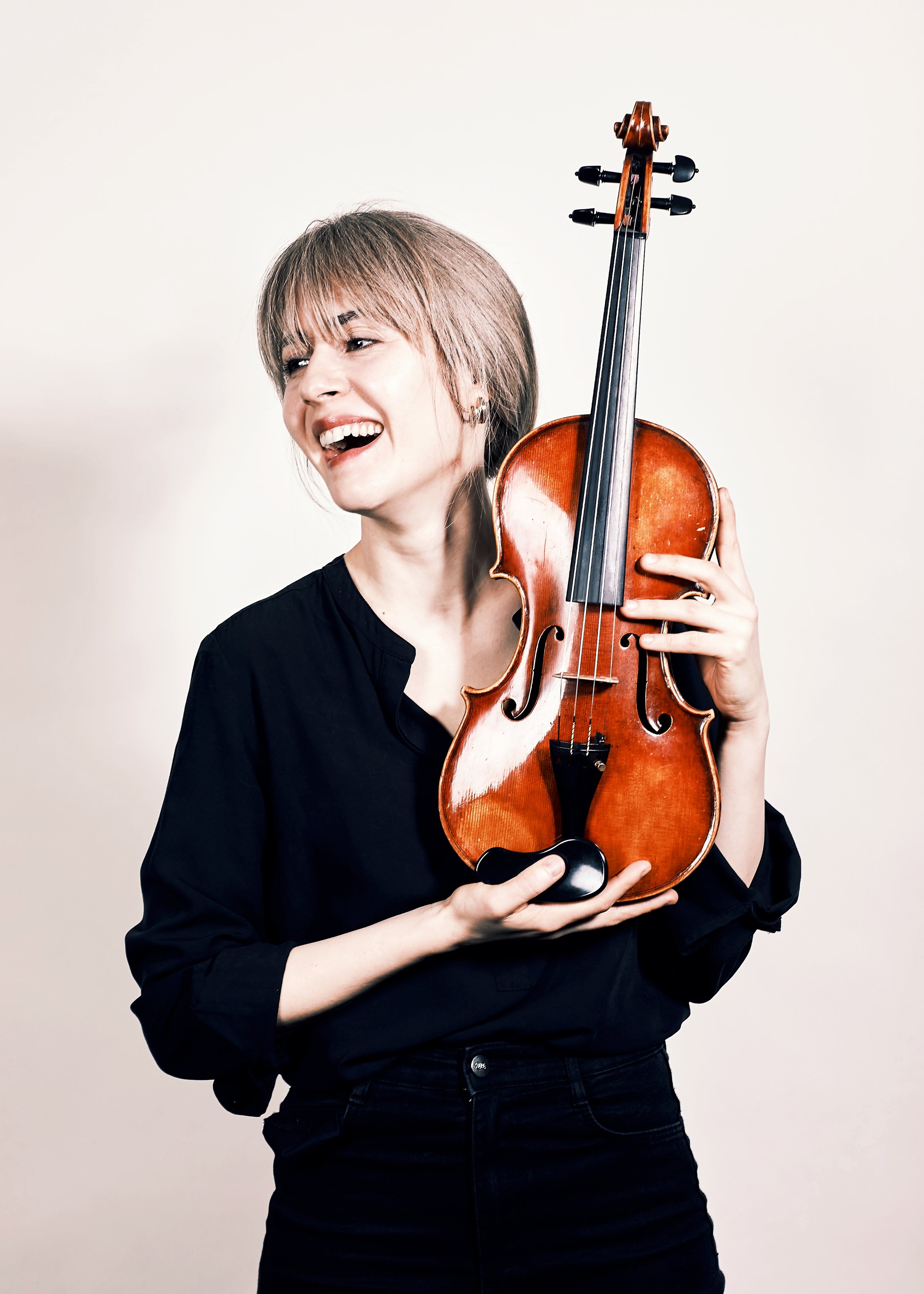 A woman, Talia Ramos, with blonde hair and dressed in black, is smiling and holding a violin close to her face against a plain white background.