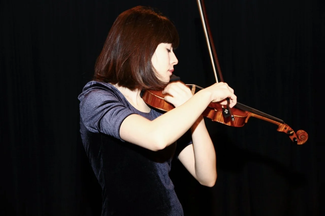 A young woman with brown hair playing a violin in a profile view, against a black background.