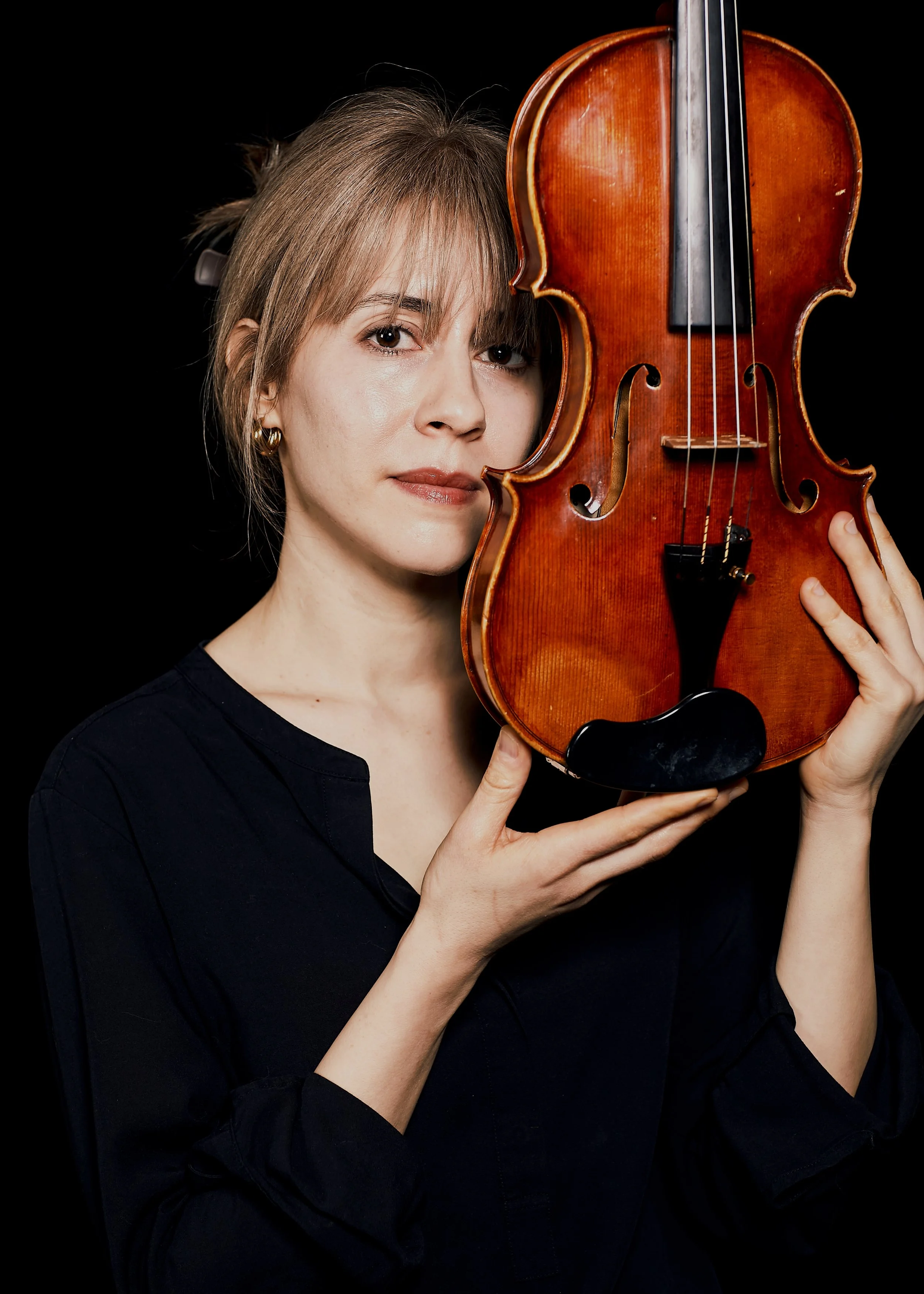 A woman, Talia Ramos, holds a violin close to her face, with a thoughtful expression, against a black background.