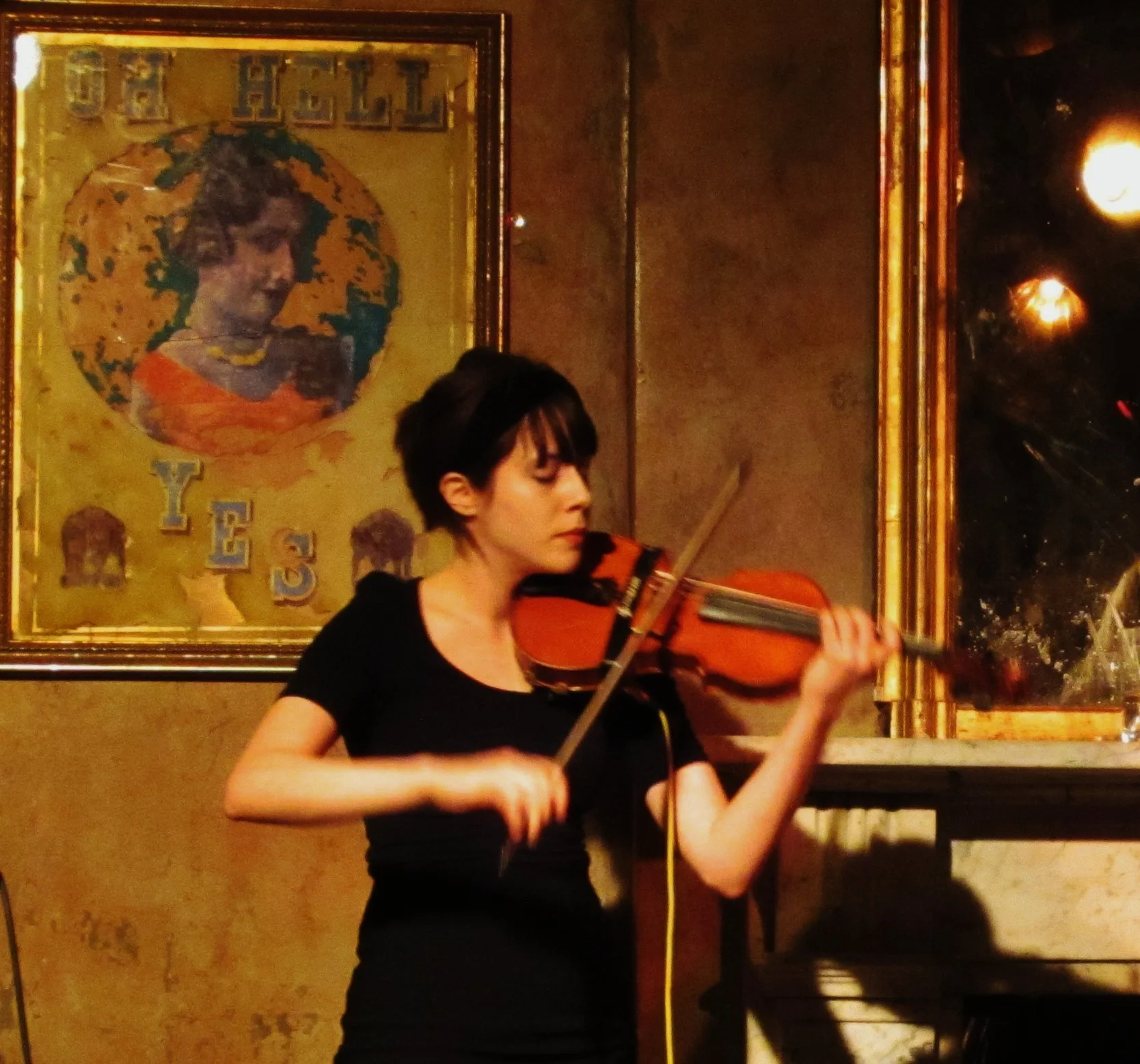 A woman, Talia Ramos, playing the violin in a London's Pub with a textured wall and framed artwork, wearing a dark top and skirt, with her hair tied back and a headband.