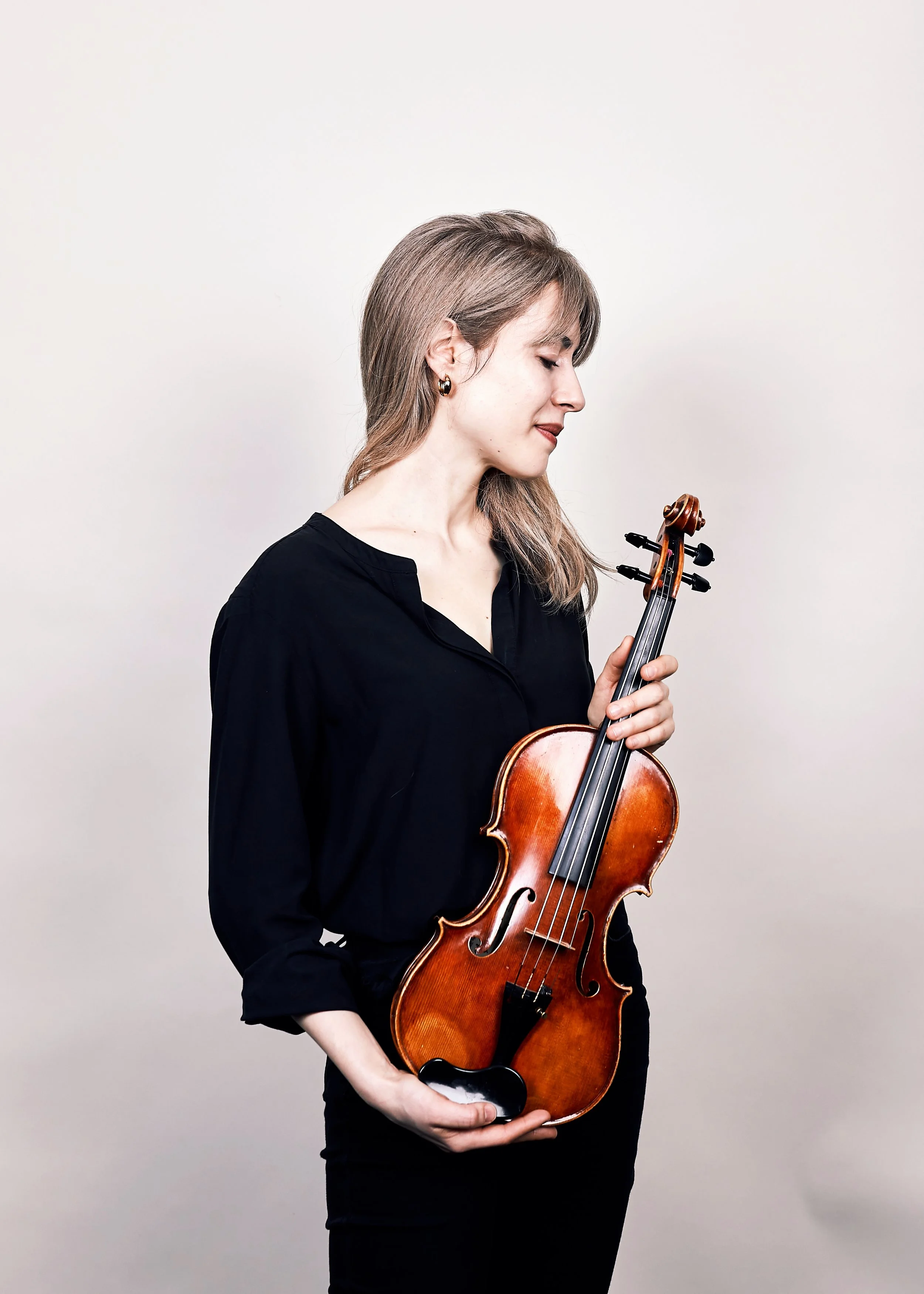 A woman, Talia Ramos, in a black shirt, holding a violin and looking at it with a peaceful expression.