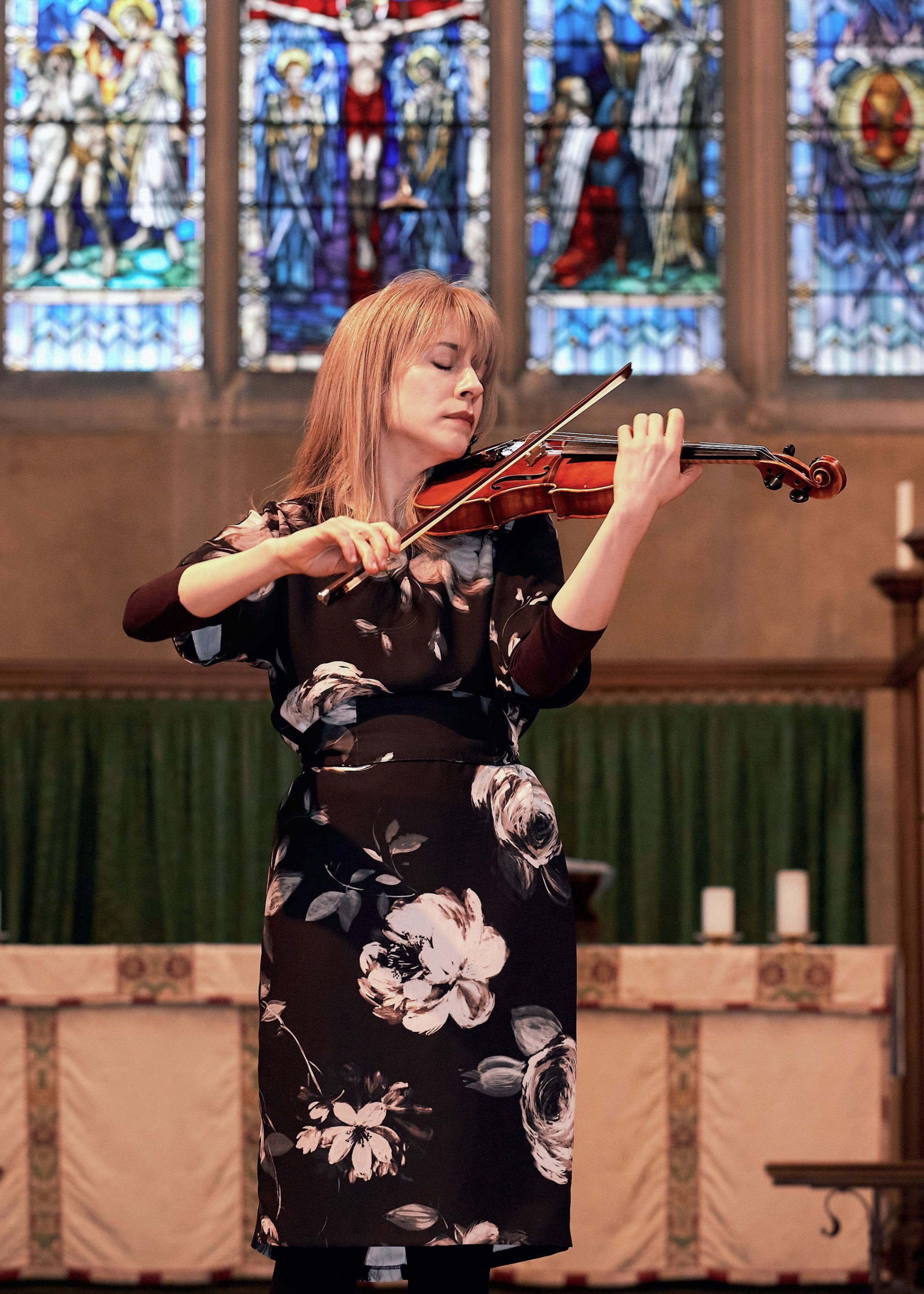 A woman, Talia Ramos, playing the violin in St Andrews Church, Muswell Hill, North London, with stained-glass windows in the background.