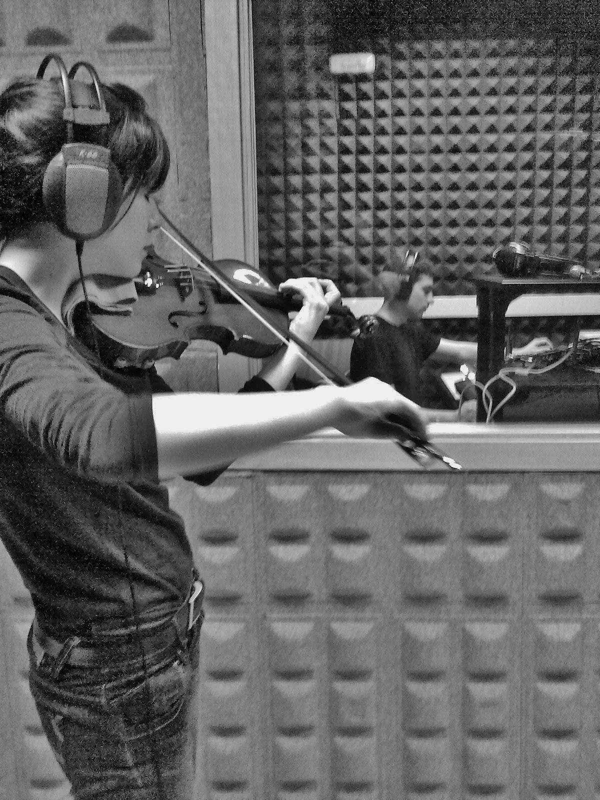 A woman in a recording studio playing a violin while wearing headphones, with the recording person working at a computer in the background.