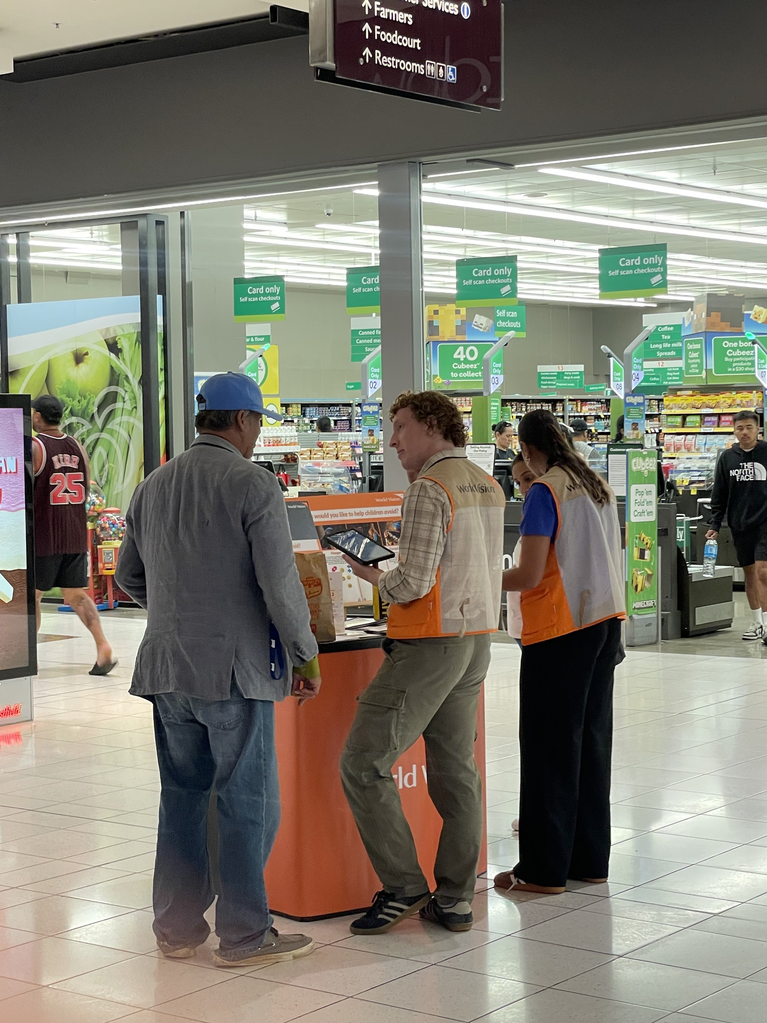 Three people at a store information desk. Two are wearing orange vests with 'WorkVision' on the back. Inside the store, signs indicate self-checkout lanes and a food court.