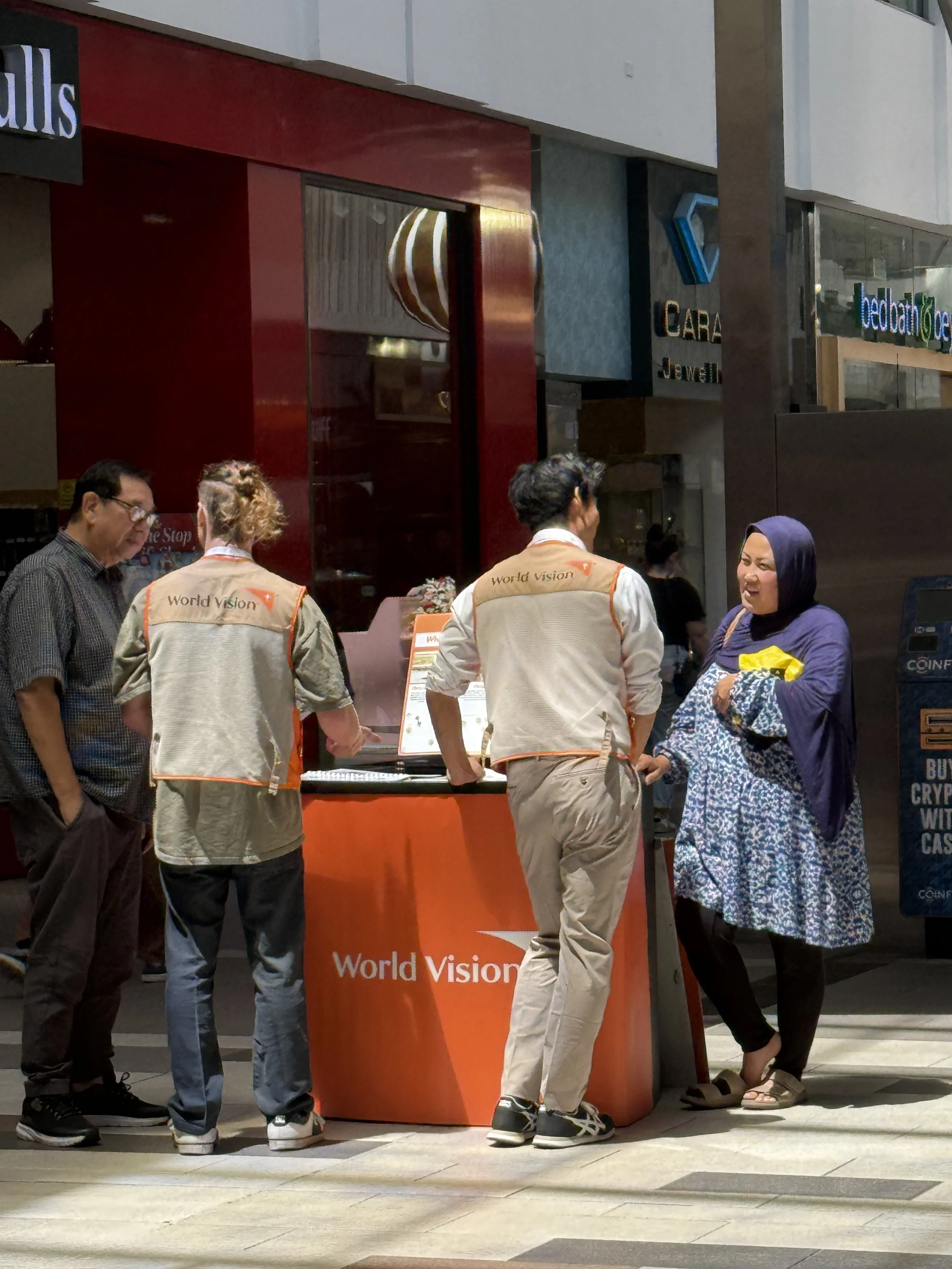 People gather around a World Vision information stand outside a storefront, engaging in conversation. The stand is orange with the World Vision logo, and the people are dressed casually, with two wearing World Vision vests.