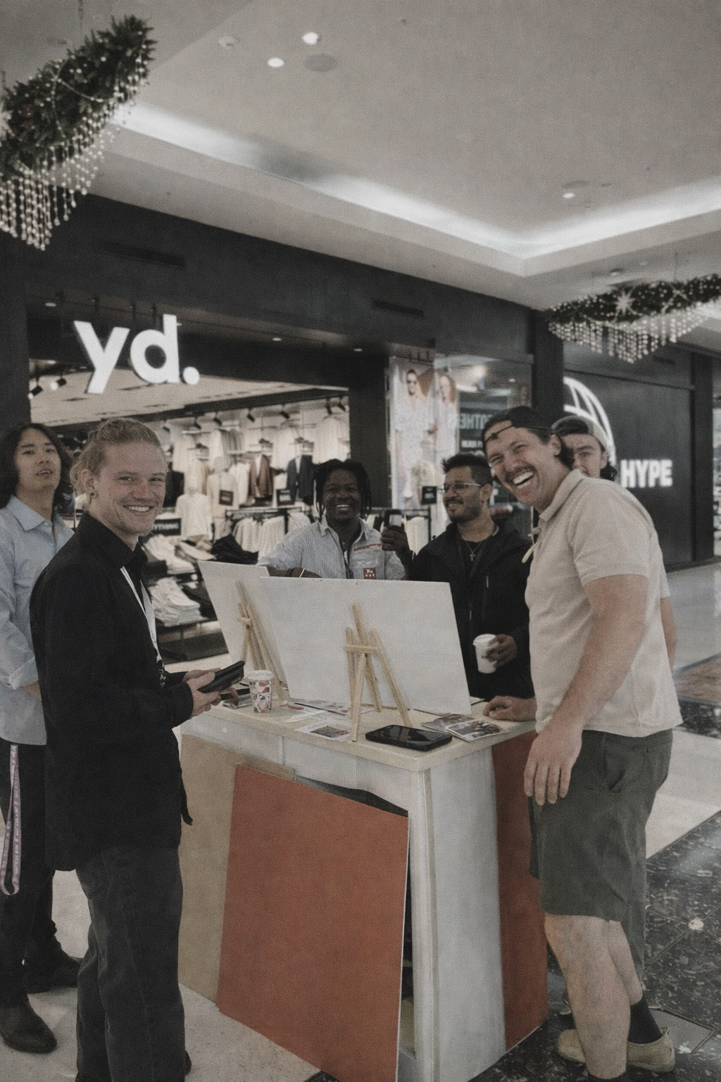 A group of happy people gathered around a booth in a shopping mall, smiling and talking, with stores and signage in the background.