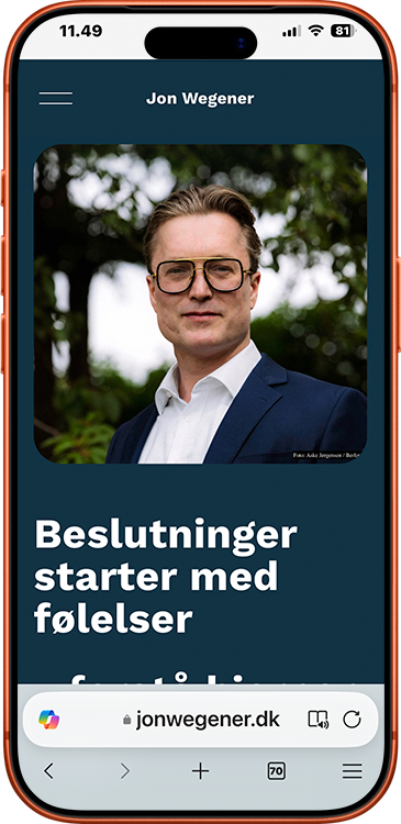 En person med brunt hår, iført sorte briller, hvid skjorte og blå blazer, står foran grønne træer.