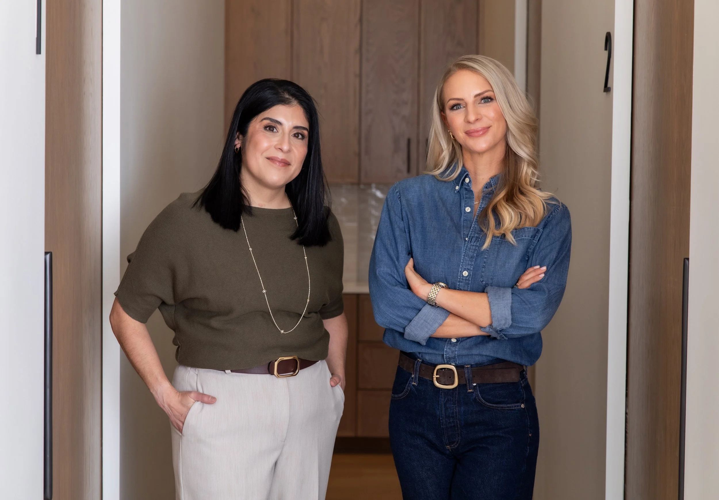 Two women standing in an indoor hallway, smiling at the camera, with wooden cabinets in the background.