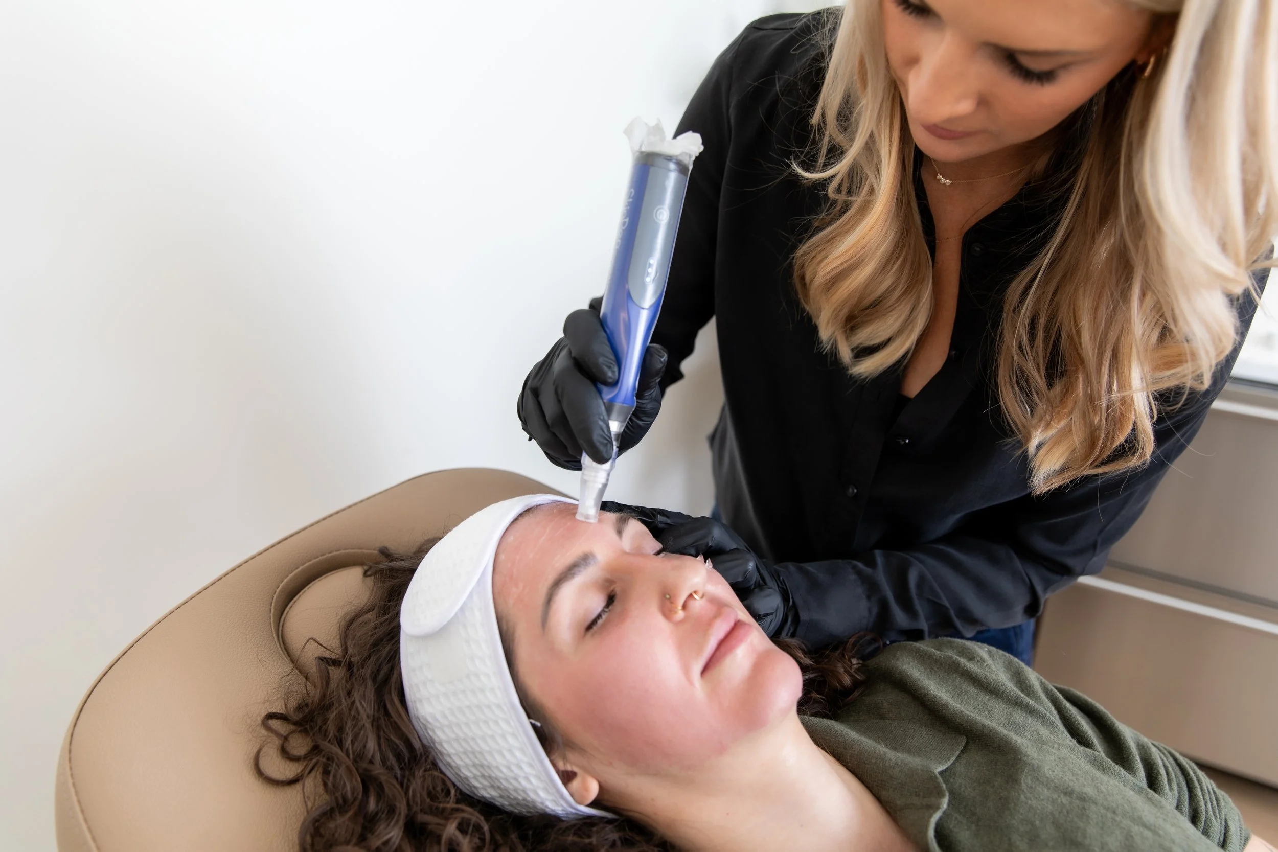 A woman receiving a facial treatment with a microcurrent device, lying on a treatment bed with a white headband, while a practitioner in black gloves administers the procedure.