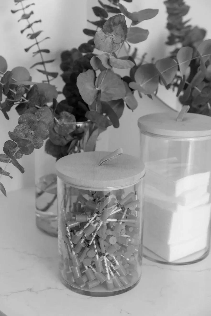 Black and white photo of a glass jar filled with syringes and another jar with cotton pads, with leafy plants in the background.