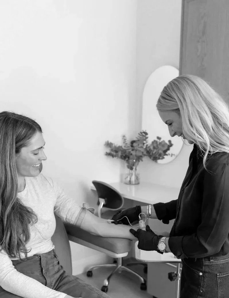 A woman is donating blood or a blood sample; she is seated and smiling while a healthcare professional, wearing gloves, prepares her arm for the procedure in a clinical setting.