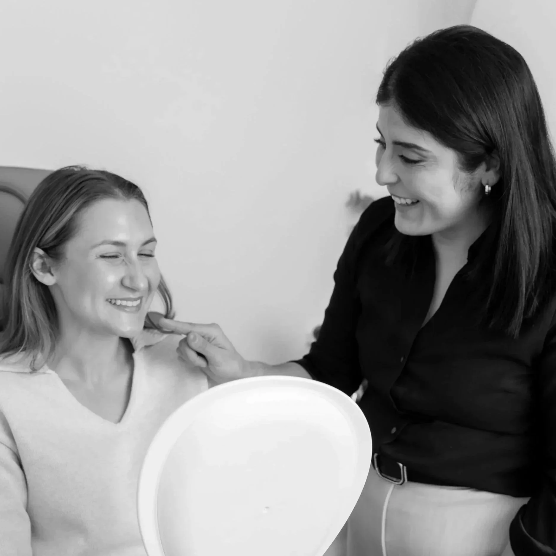 A woman with shoulder-length hair smiling as another woman with dark hair applies makeup to her face, with a circular mirror on a stand in front of them, in a casual indoor setting.
