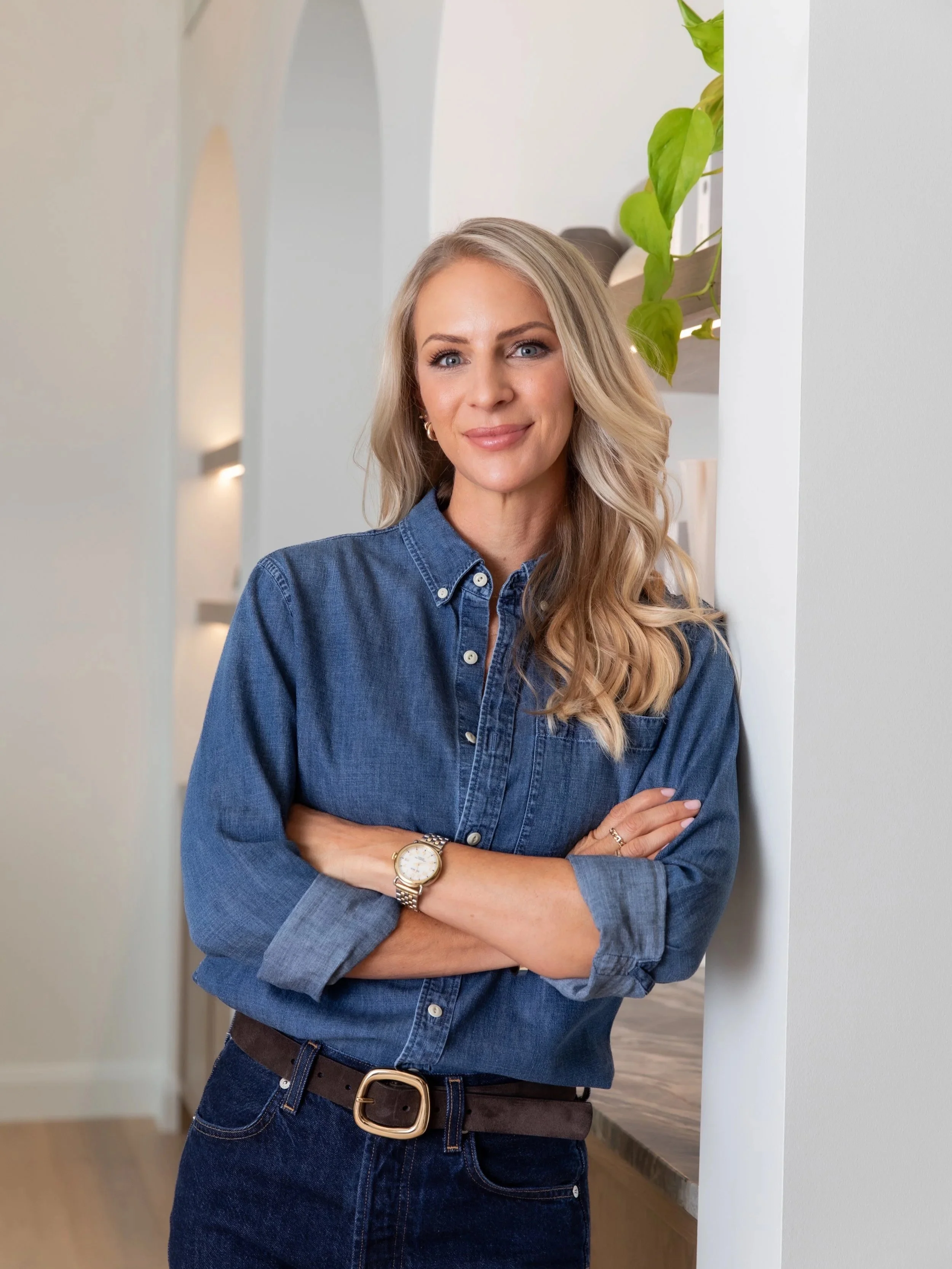 A woman with long blonde hair wearing a denim shirt and dark jeans, standing with crossed arms in a modern, well-lit interior with a white wall and a leafy green plant behind her.